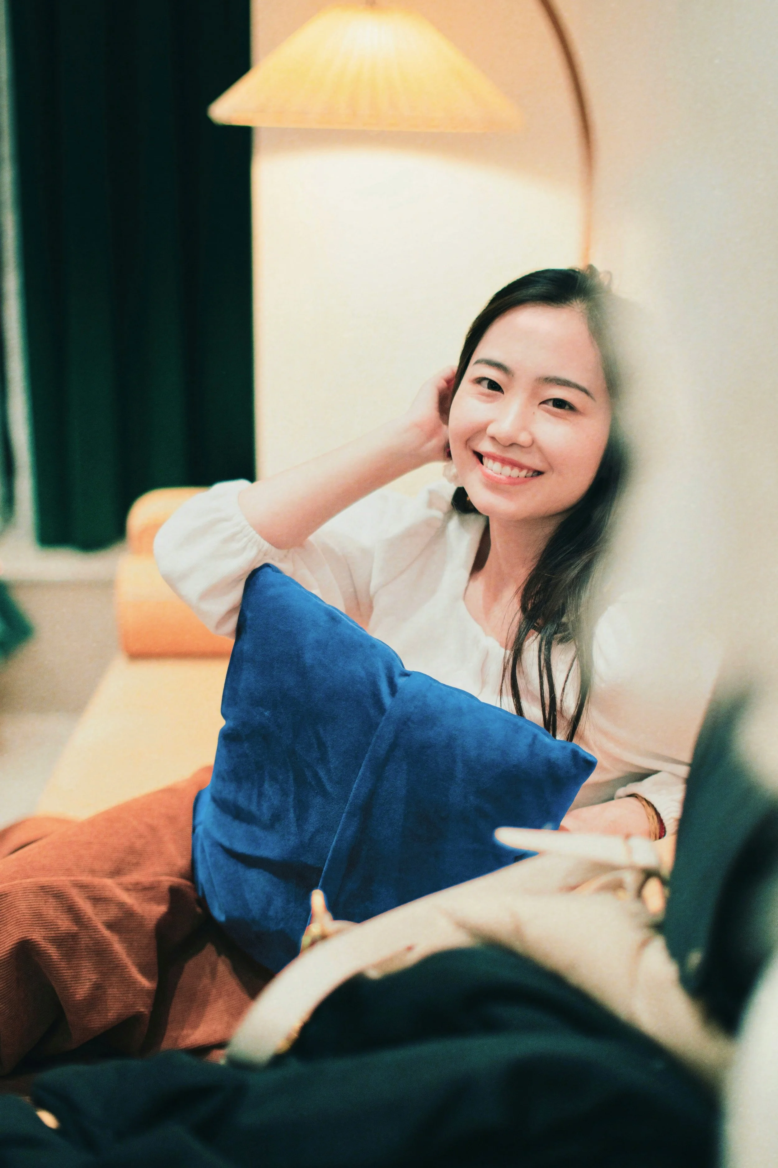 An Asian woman sits on a couch and smiles at the camera, she's holding a blue pillow