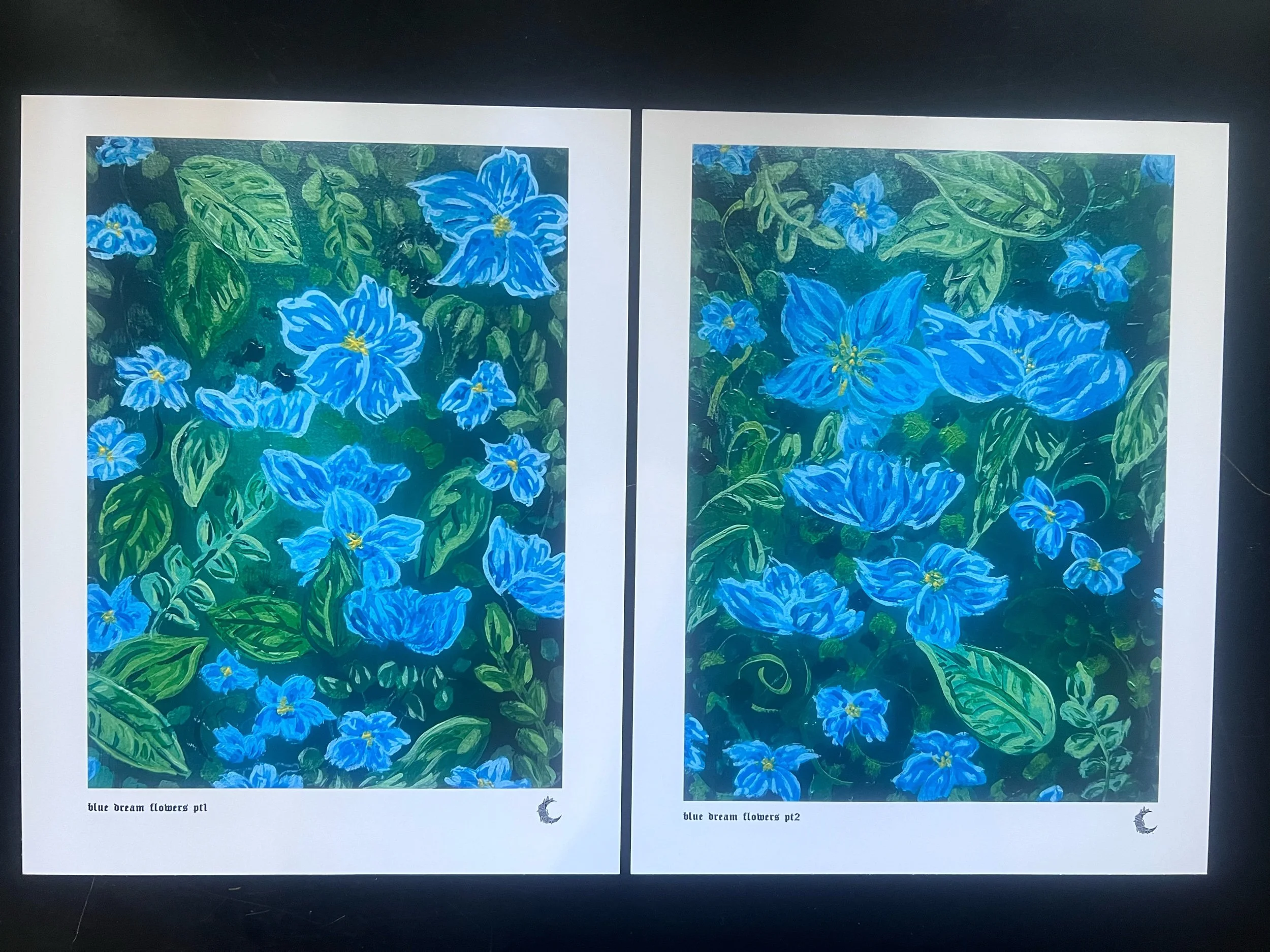 A colorful floral illustration of blue flowers with green leaves on dark gren background, printed on photo paper with the tites at the bottom left. There are dried roses and various small rocks around the paper.