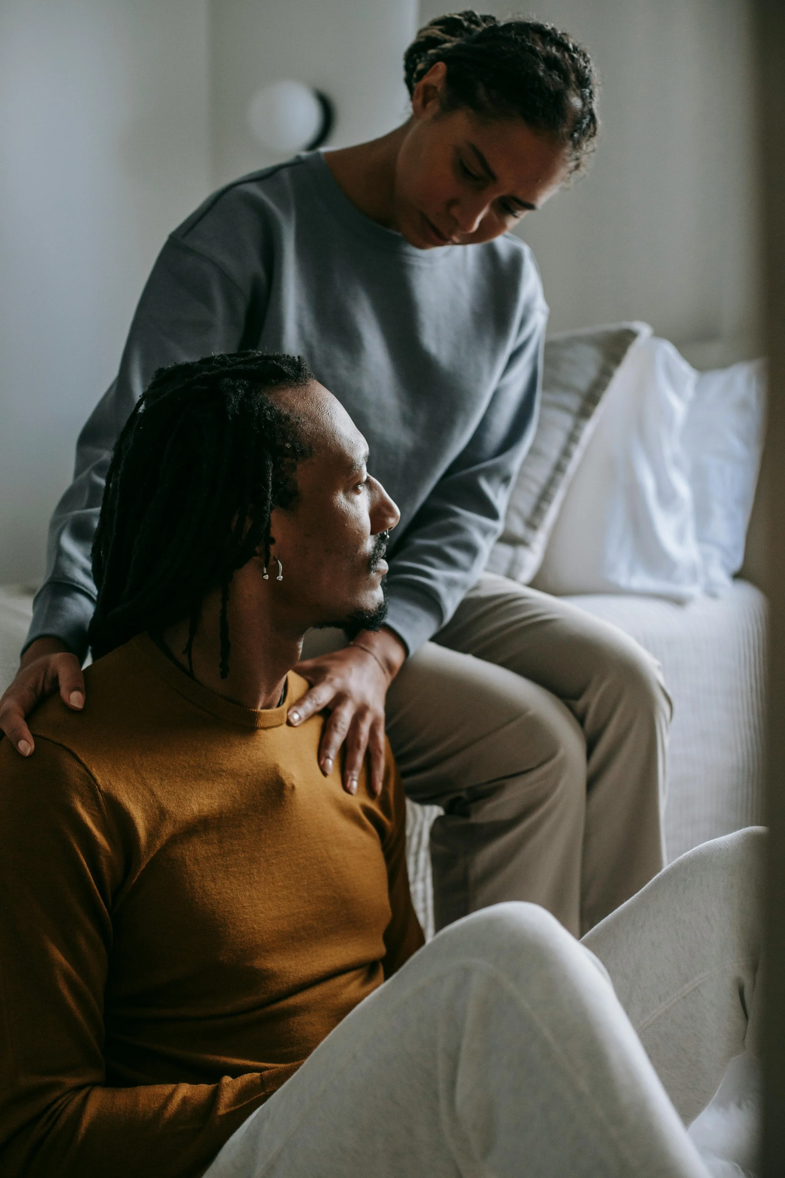 A woman sits on a bed with her hand on a man's shoulder as he sits on the floor, both appearing serious in a softly lit room, the weight of betrayal evident in their expressions.