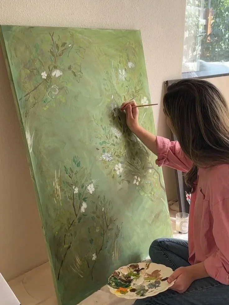 A woman sitting cross-legged, painting a large green canvas with white flowers and foliage, using a small brush and a palette of mixed paints.