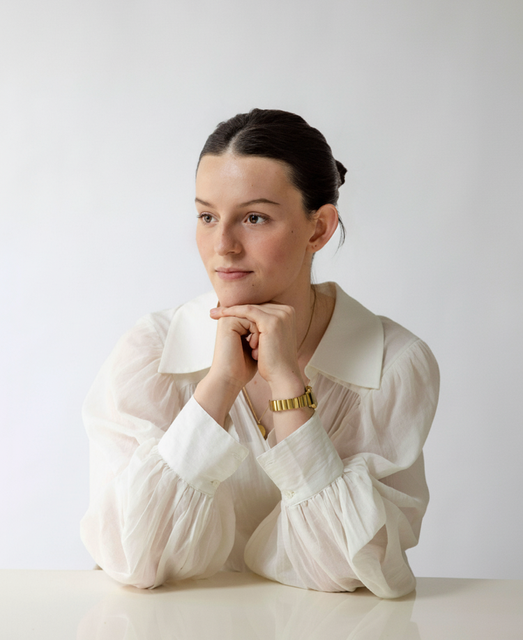 A young woman with dark hair in a bun, wearing a white blouse and a gold watch, sitting with her chin resting on her hands, looking thoughtfully to the side against a plain white background.
