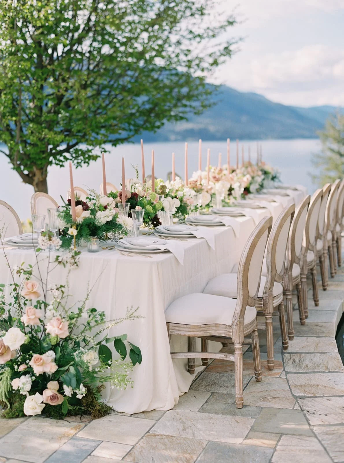 A stunning formal outdoor wedding reception table overlooking a lake. The table is filled with lush Twig & Bloom flower centerpieces. There are tall taper candles between plenty of fresh spring wedding floral centerpieces.