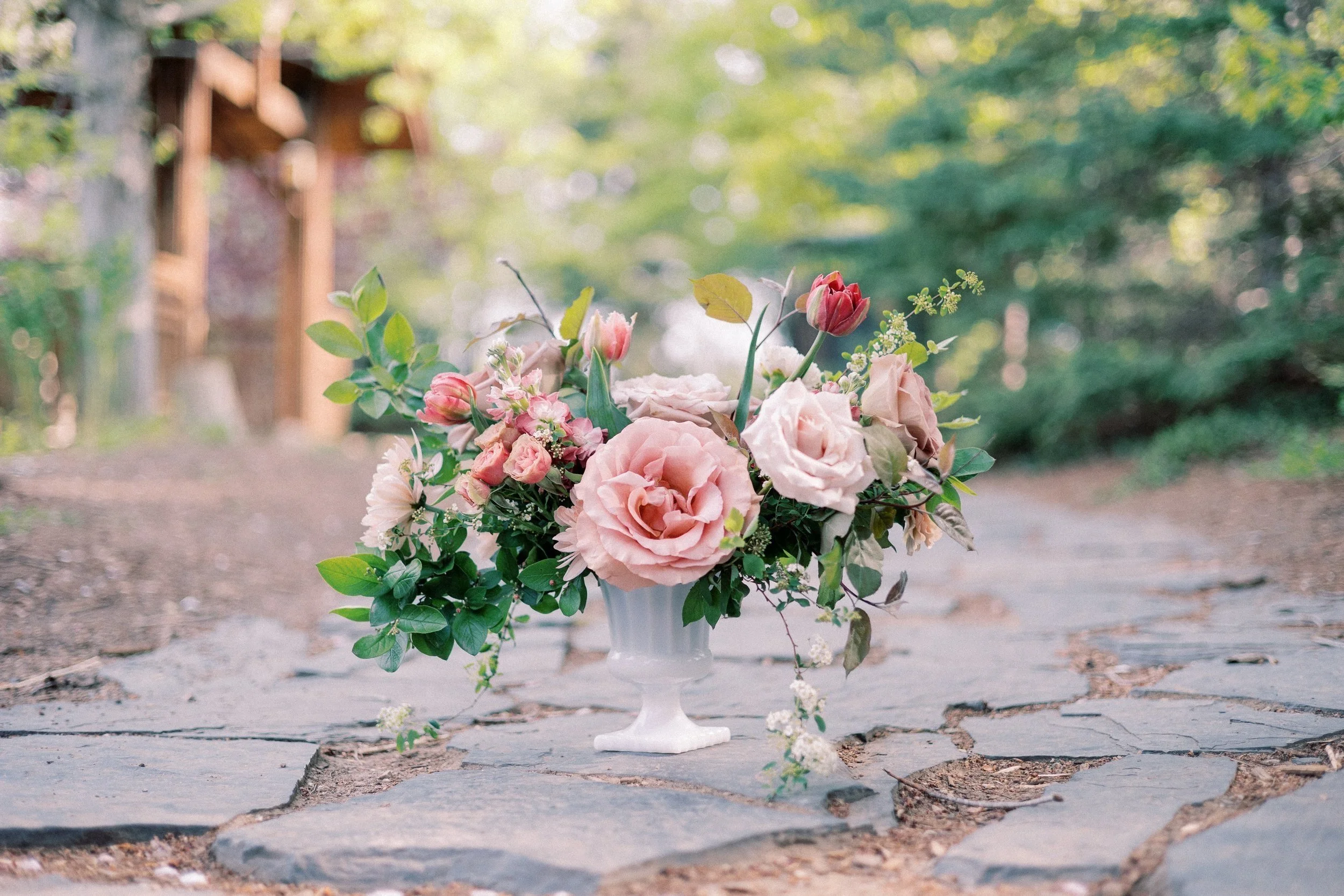 A Twig & Bloom arrangement of various flowers and greenery in a white pedestal vase on a stone pathway in an outdoor garden setting.