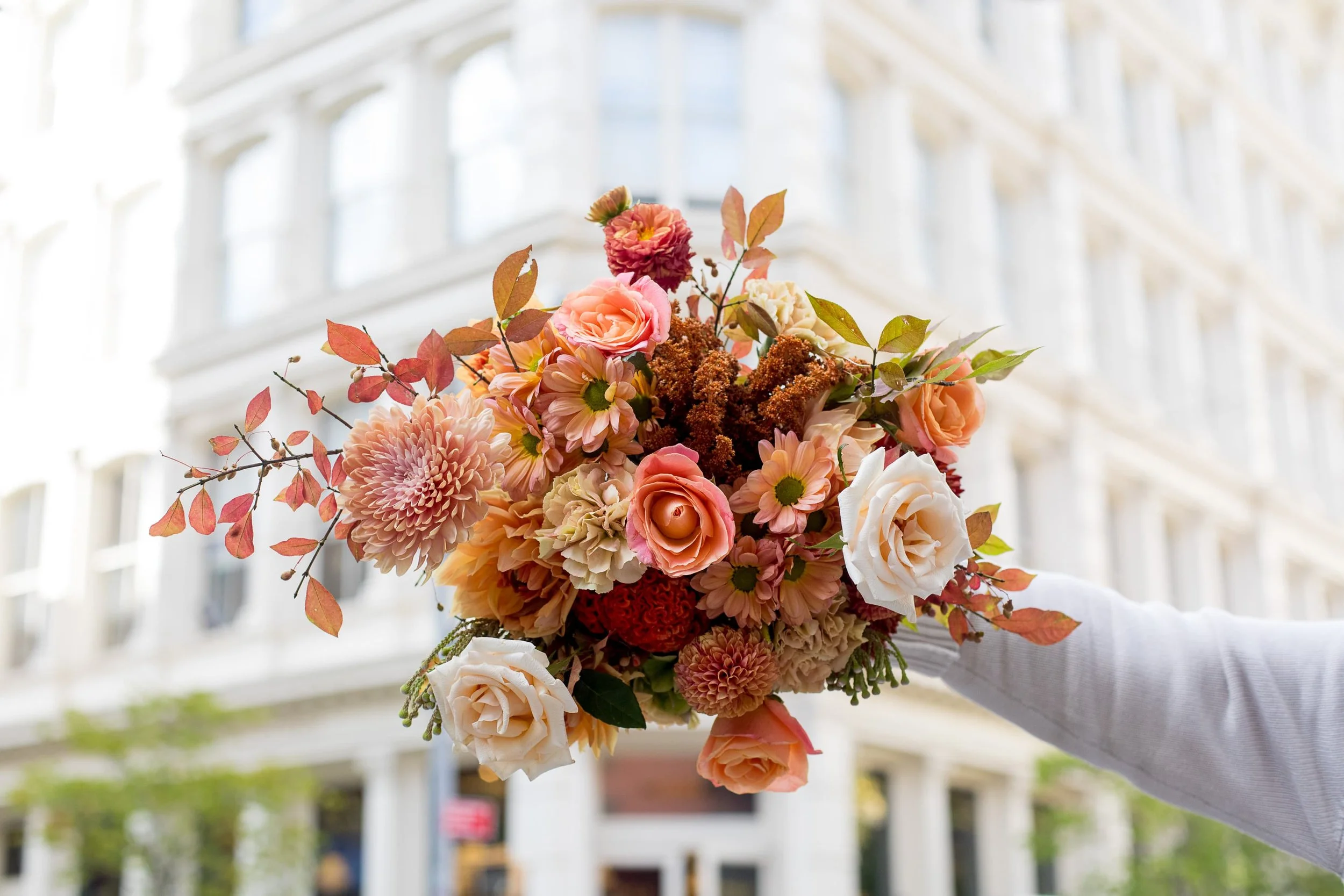 A person holding a Twig & Bloom colorful bouquet of flowers, including roses and daisies, indoors with a bright, industrial-style window and building in the background.