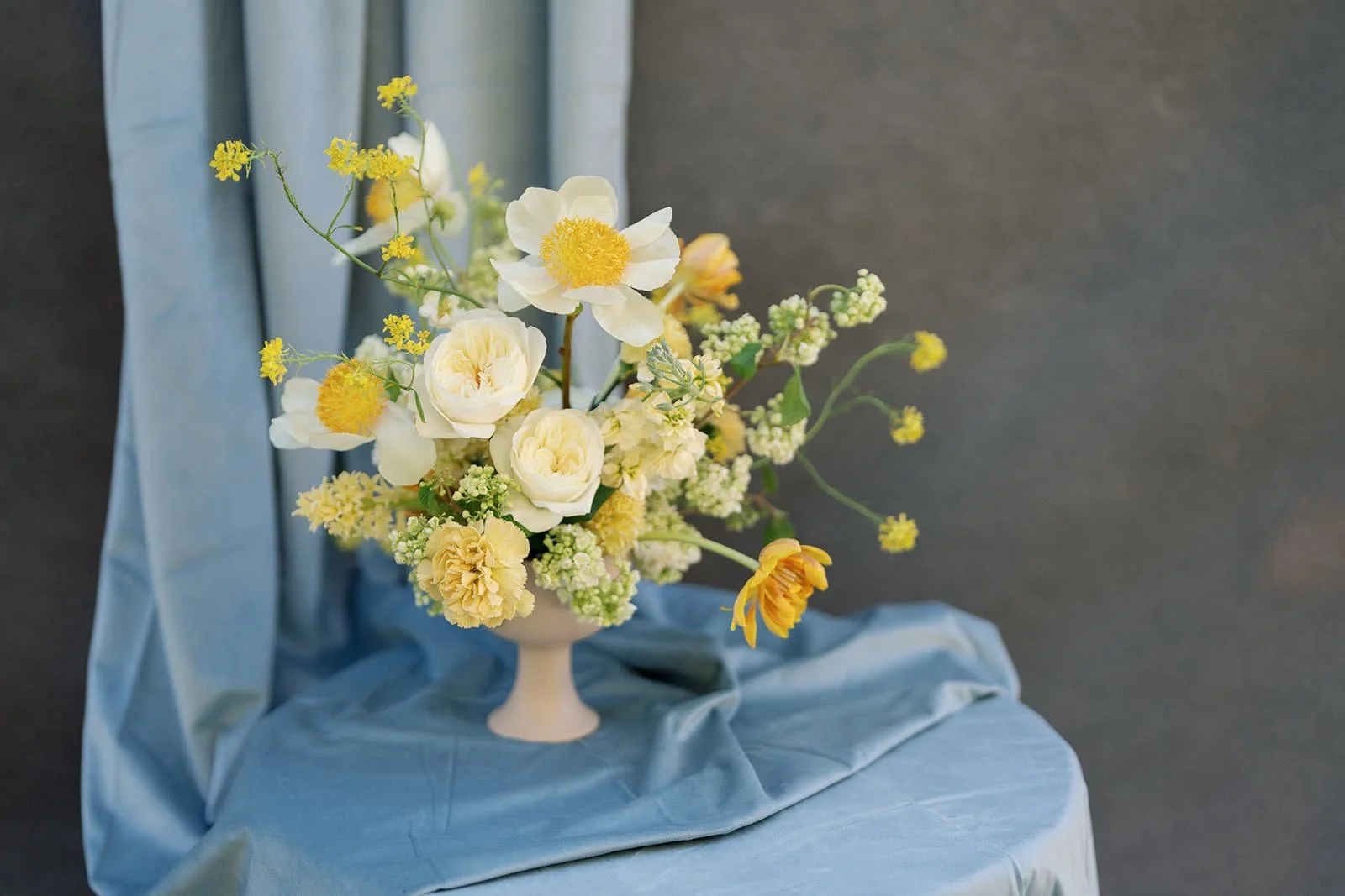 An arrangement of yellow flowers in a cream pedestal vase placed on a blue fabric-covered surface with a blue fabric backdrop.
