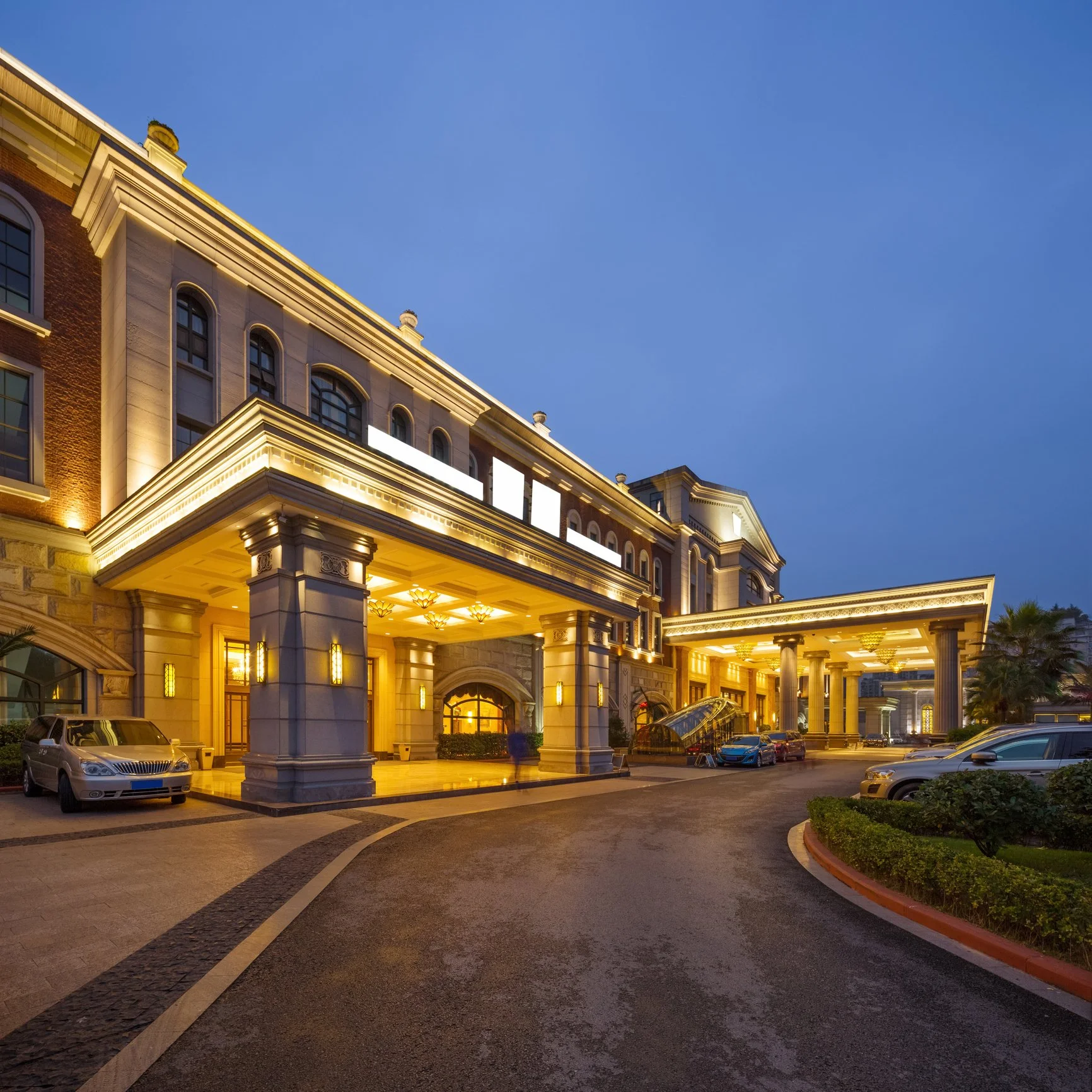 Luxury hotel entrance with illuminated columns and a driveway lined with parked cars, during twilight.