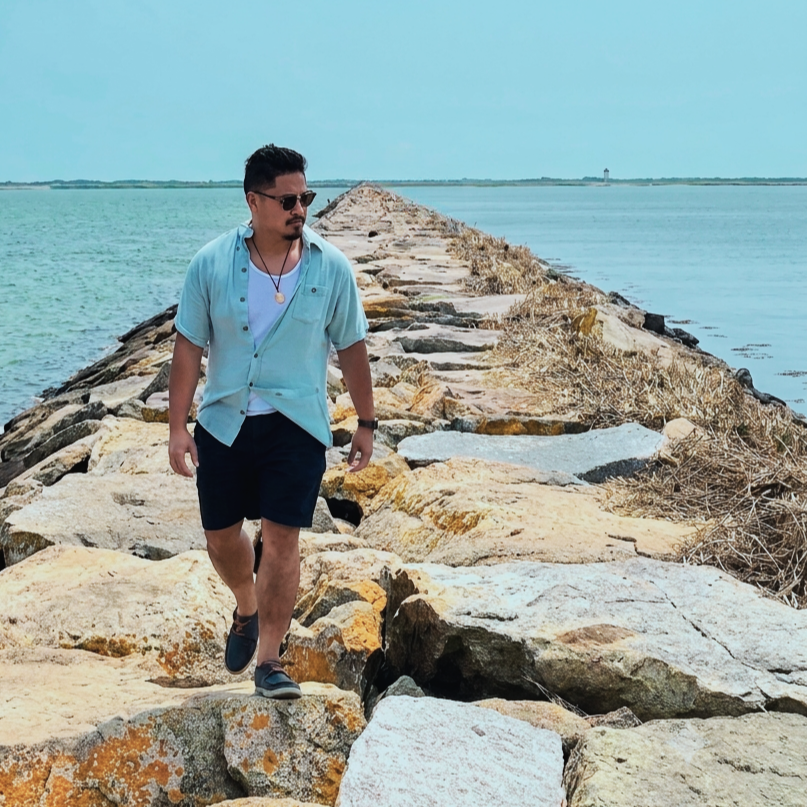Man walking on a rocky pier near a body of water, wearing sunglasses, a light blue shirt, black shorts, and sneakers, with a distant lighthouse in the background.