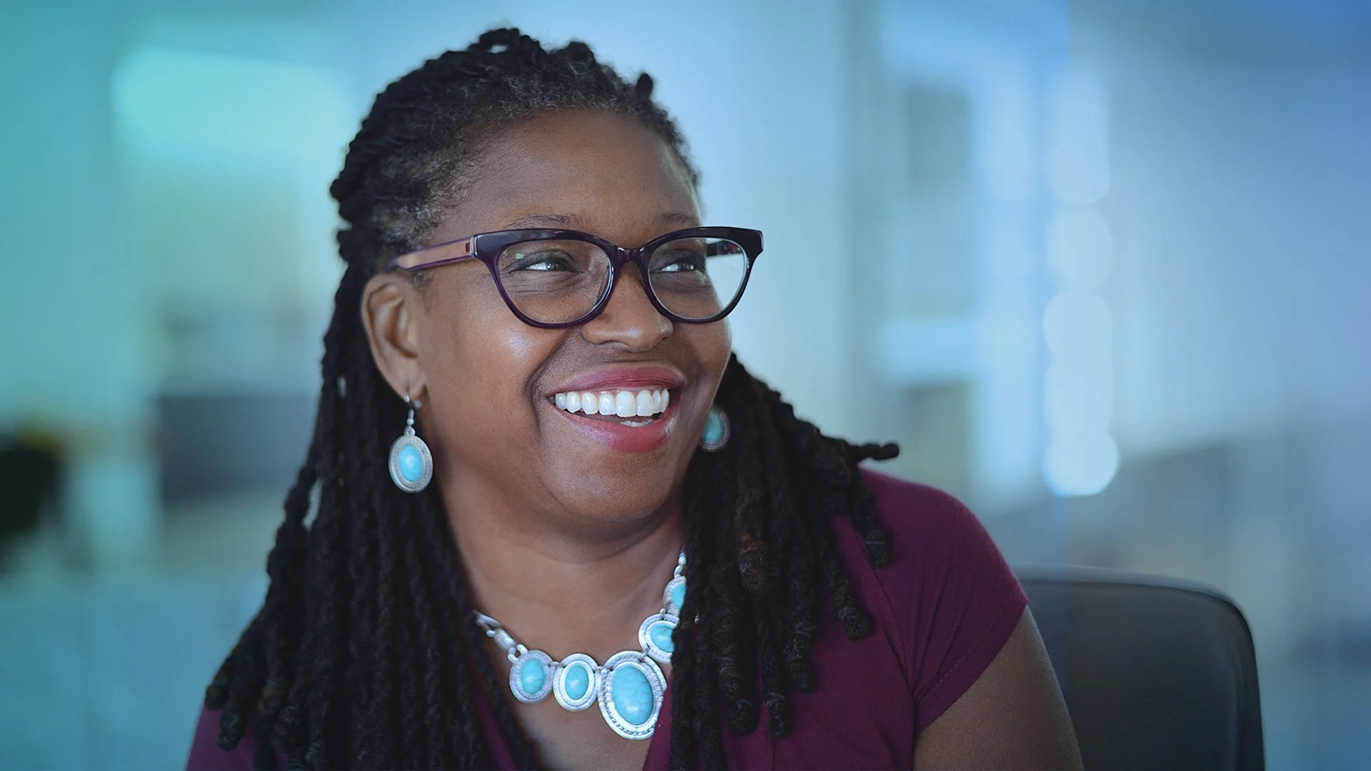A smiling African American woman with glasses, wearing a purple top and turquoise jewelry, in a bright indoor setting.