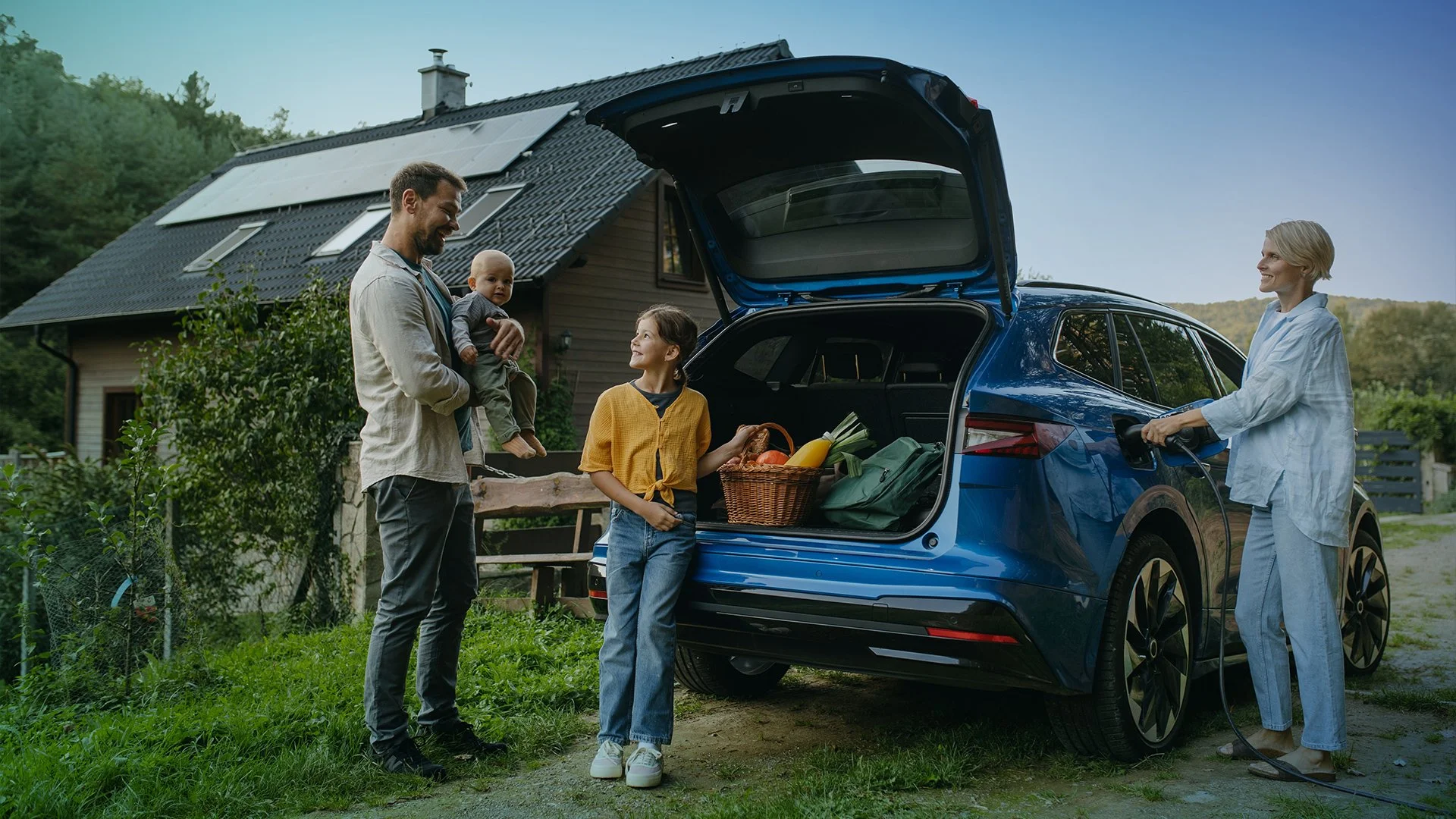 A family is unloading groceries from an electric car in their driveway, with a house and trees in the background.