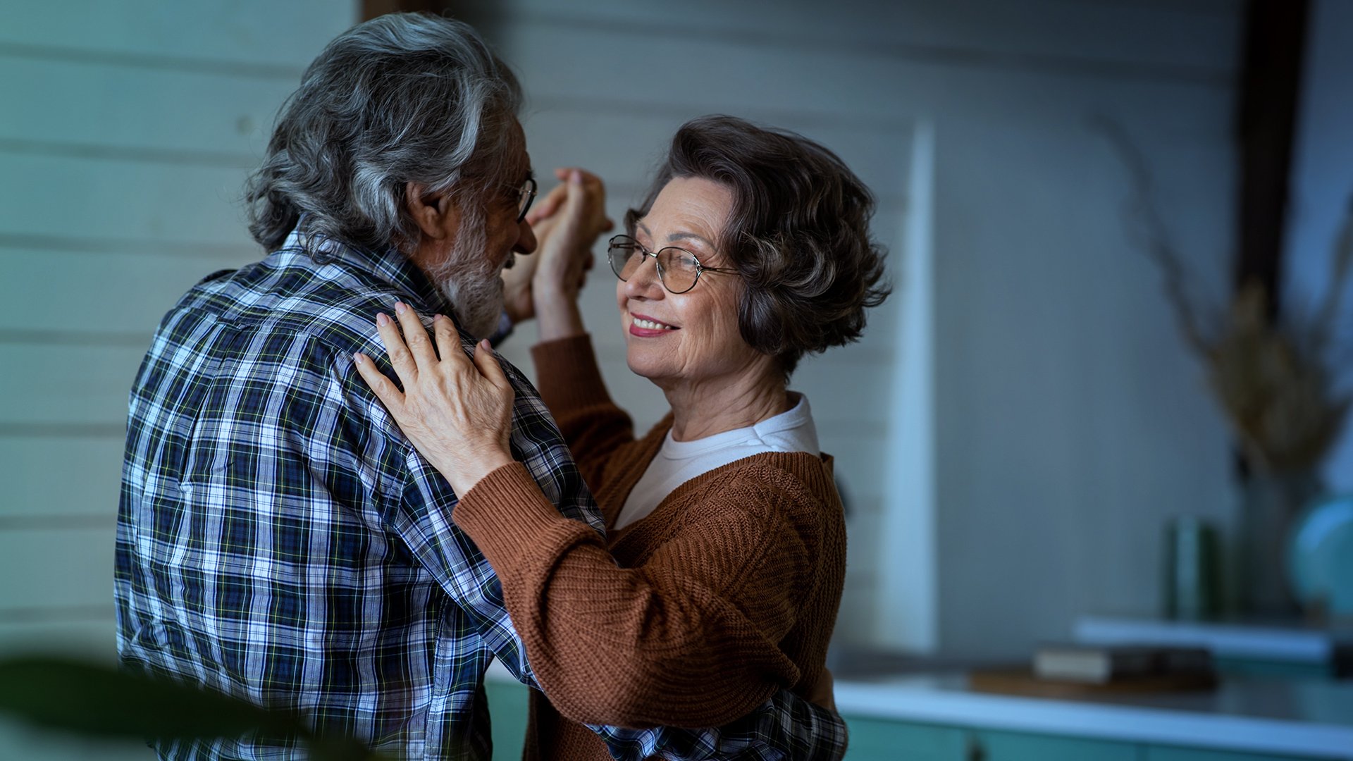 An elderly couple dancing closely together inside a home, smiling and looking at each other.