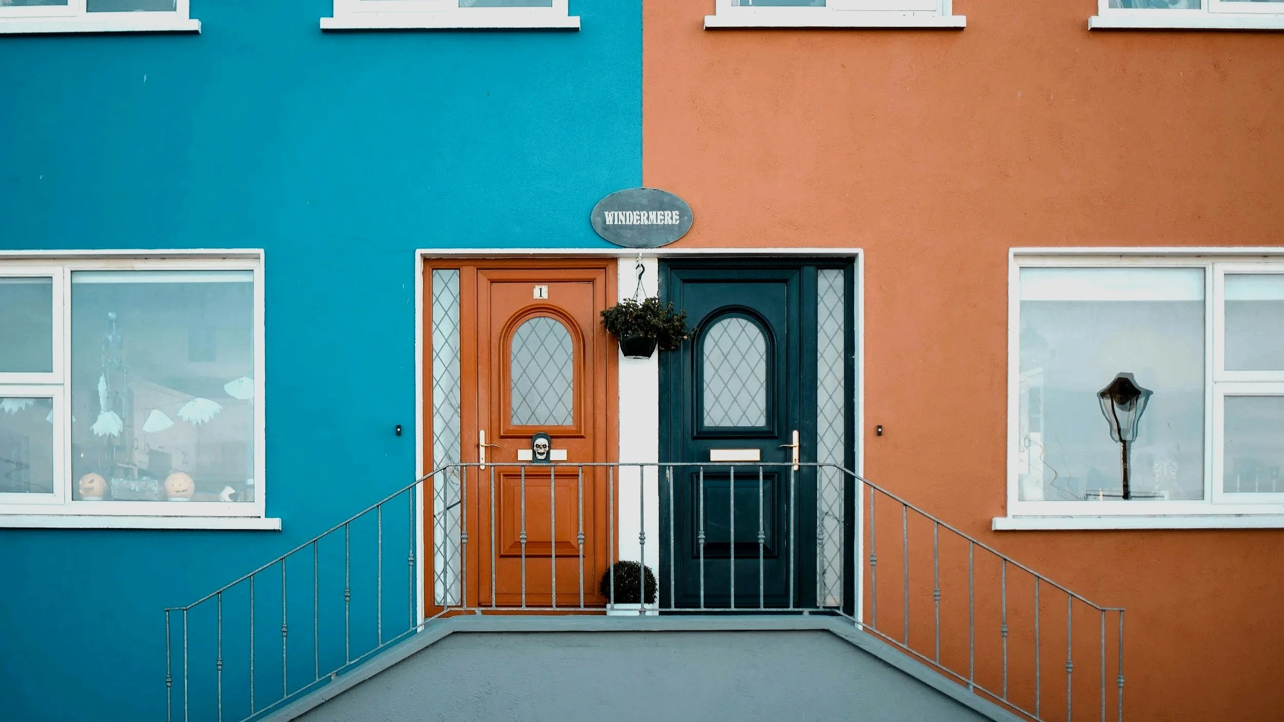 Colorful apartment building with two doors, one orange and one dark green, framed by orange and blue walls, with a sign that says ''Windermere'' above the doors, and decorative window and street lamp visible through the windows.