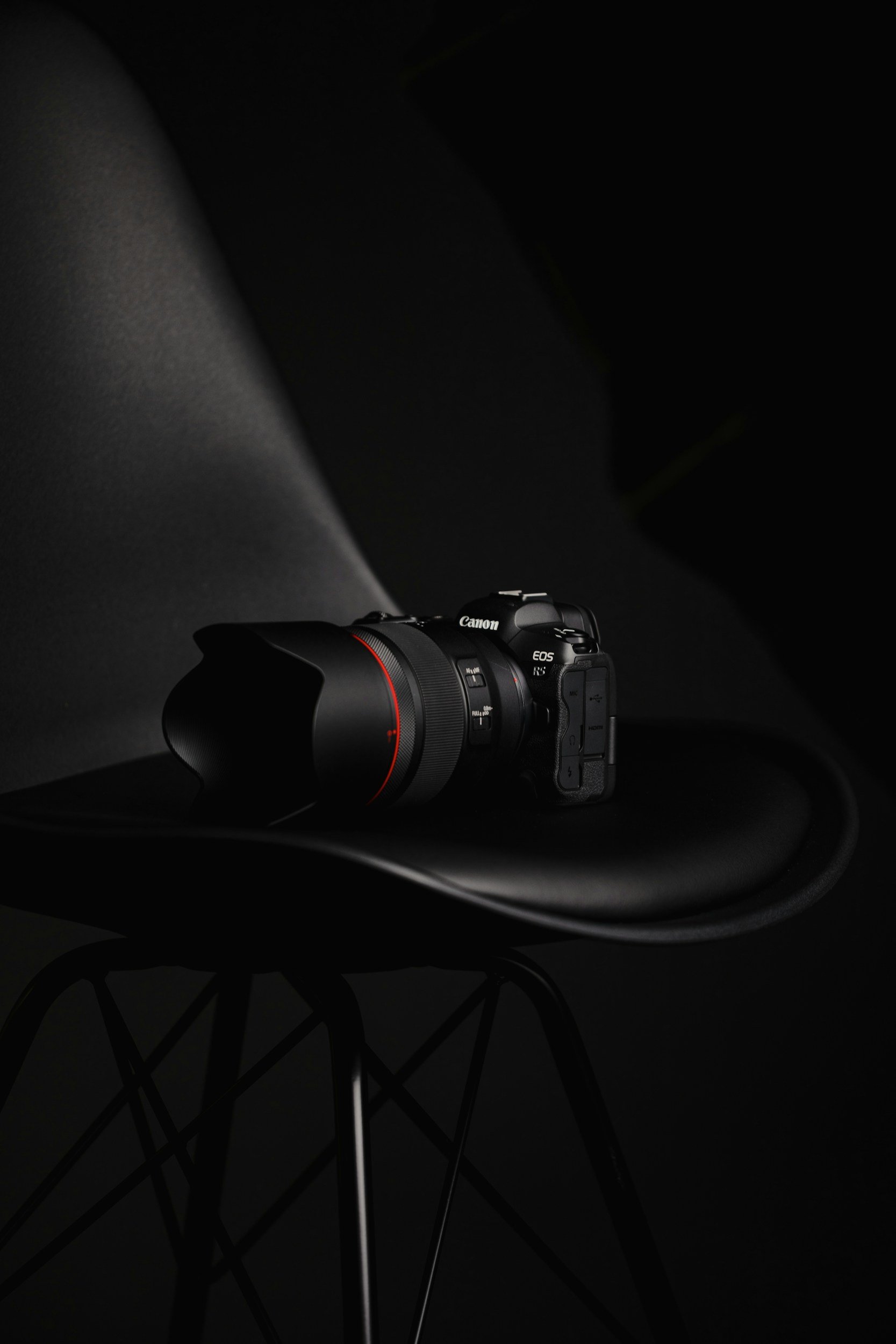 A Canon EOS R5 camera with a large lens resting on a black chair against a dark background.
