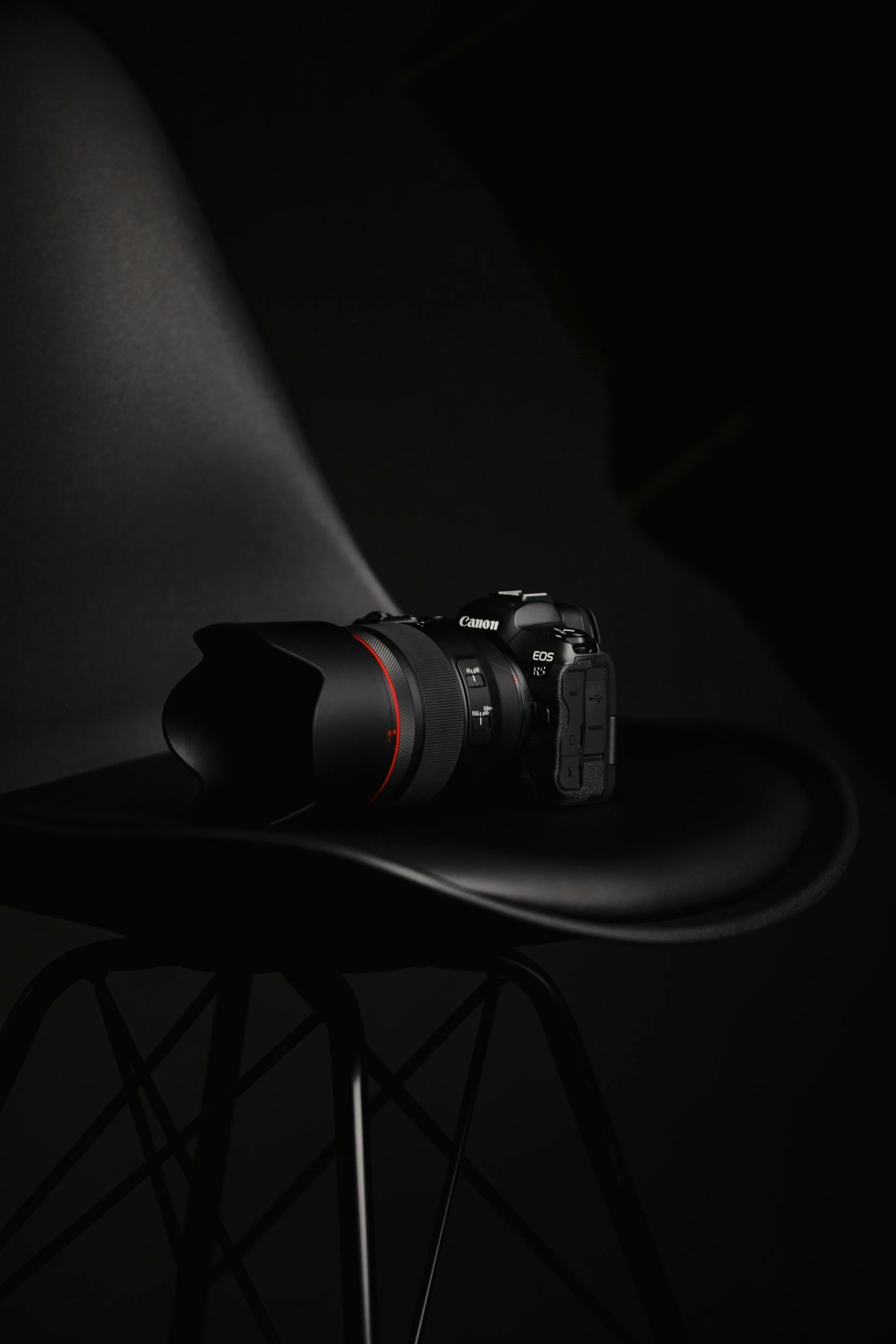 A Canon EOS R5 camera with a large lens mounted on a black chair against a dark background.