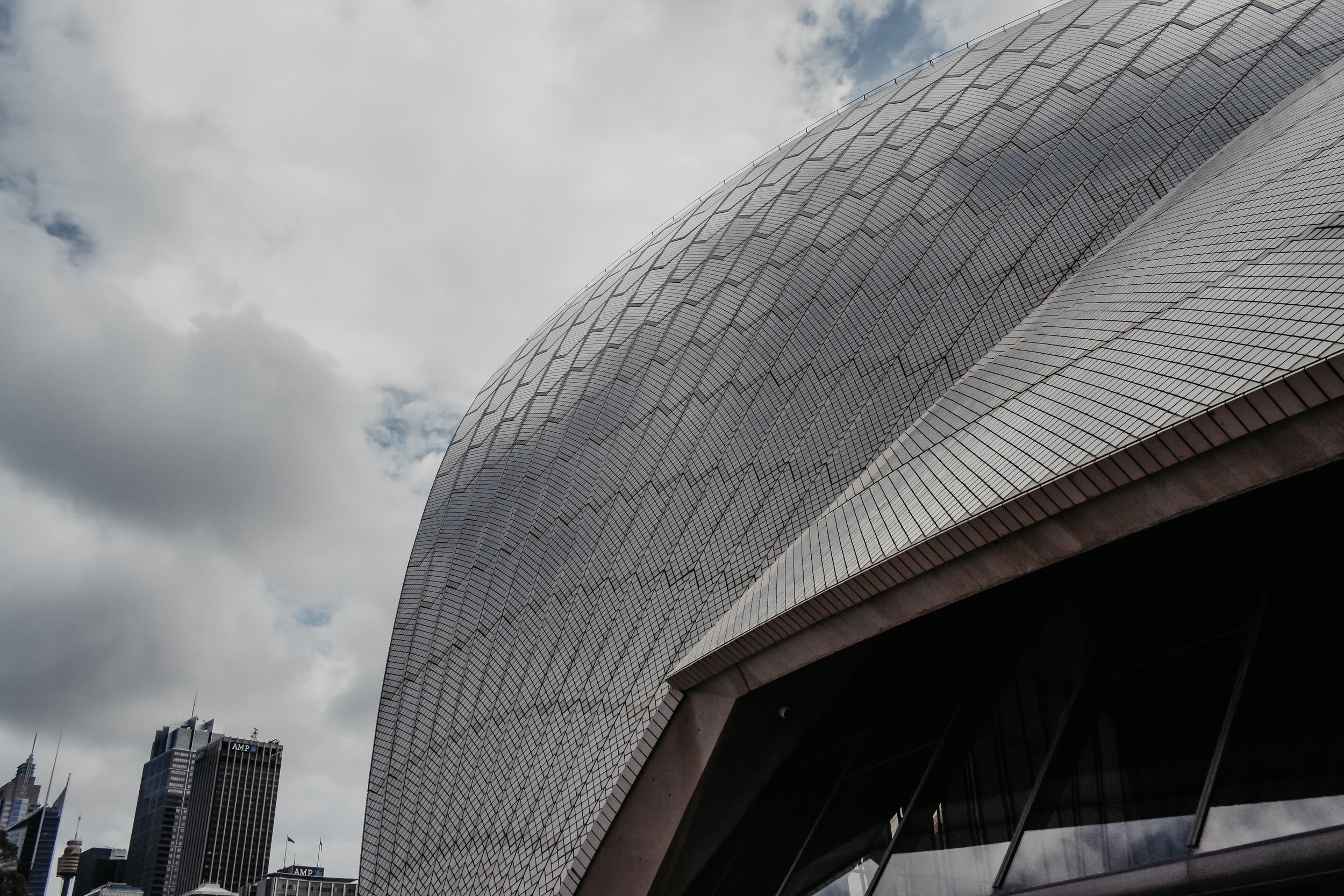 Close-up of a modern glass building with a curved, reflective surface and geometric patterns, with a city skyline and cloudy sky in the background.