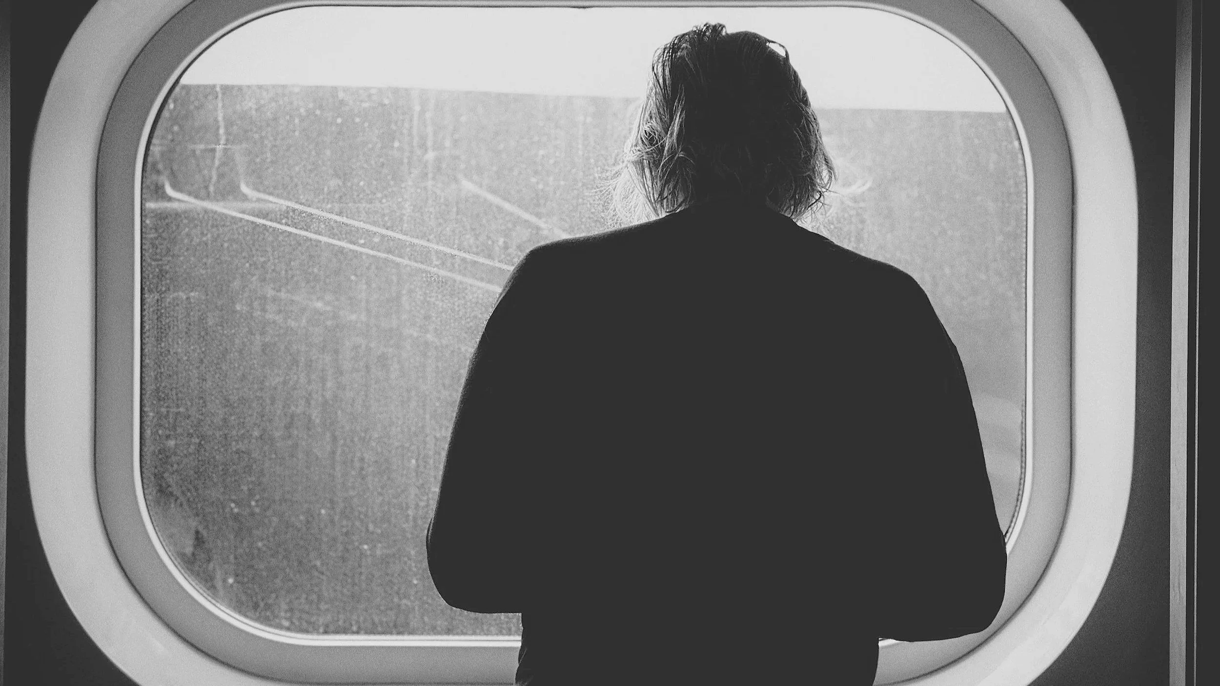 A person with medium-length hair viewed from behind, standing in front of an airplane window, looking outside on a cloudy day.