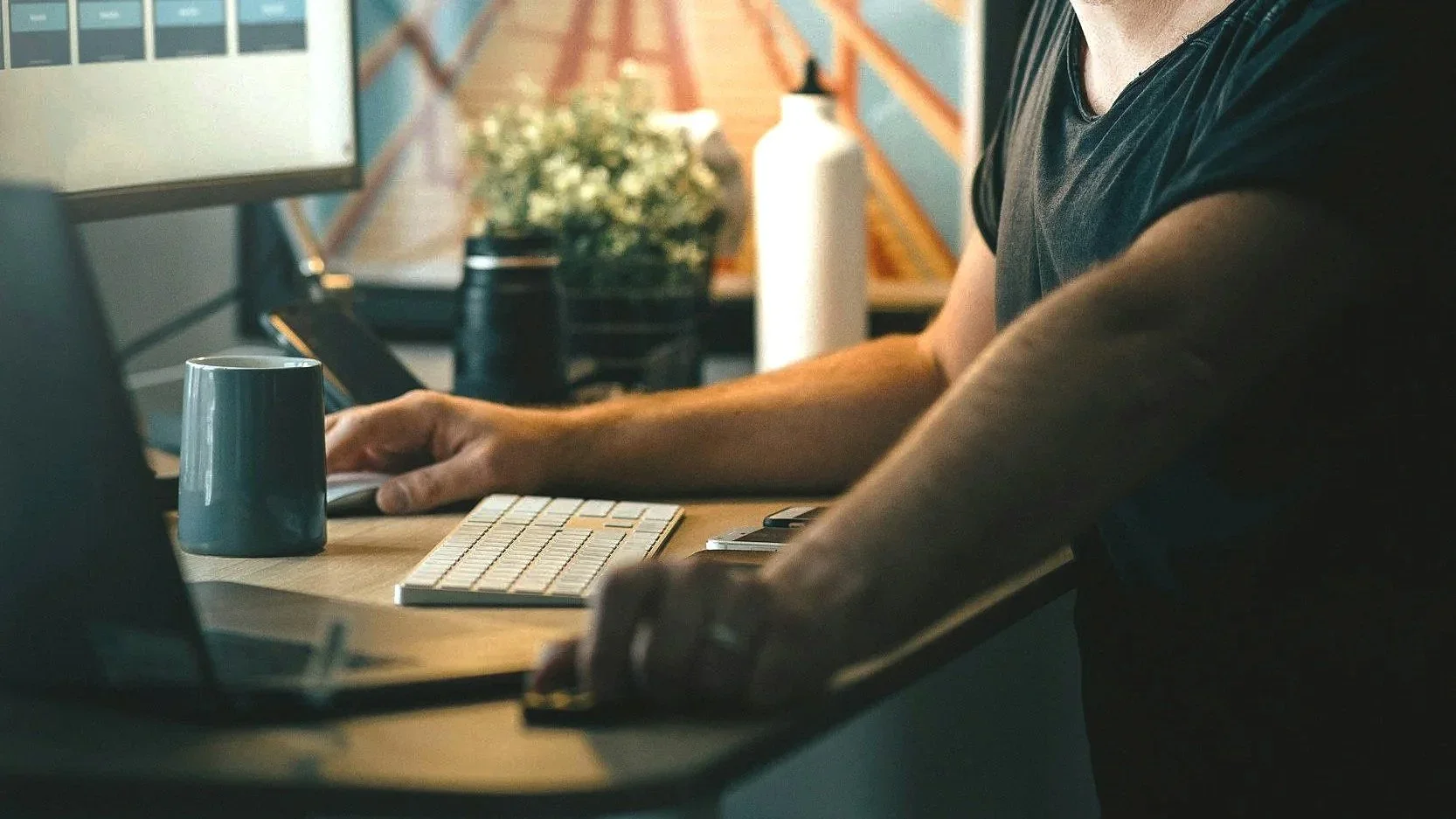 Person working at a desk with a computer, keyboard, mug, and various office items, in a modern workspace.