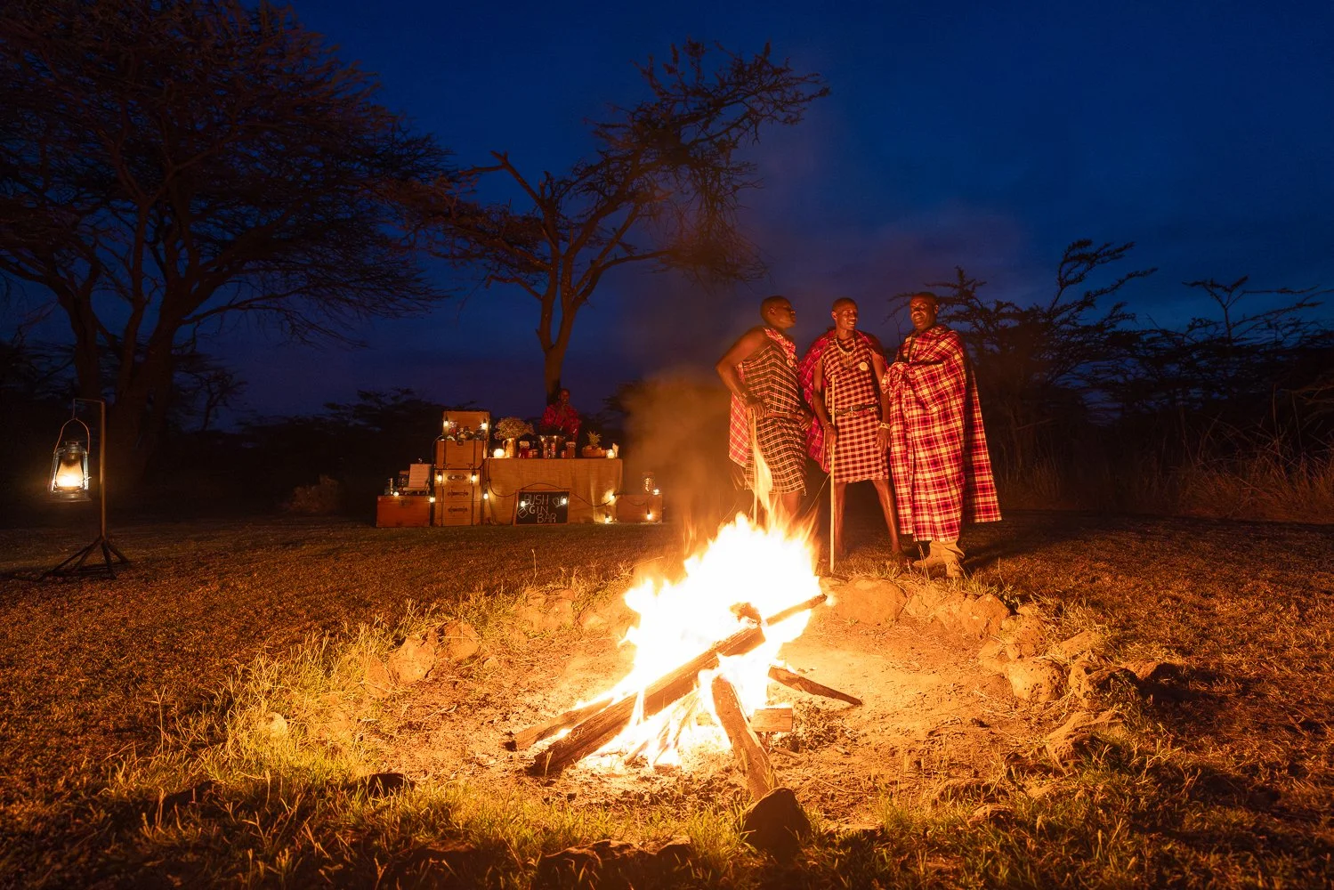 Three Maasai men in traditional attire standing around a campfire at night with trees in the background and a decorated setup with food and drinks in the distance.