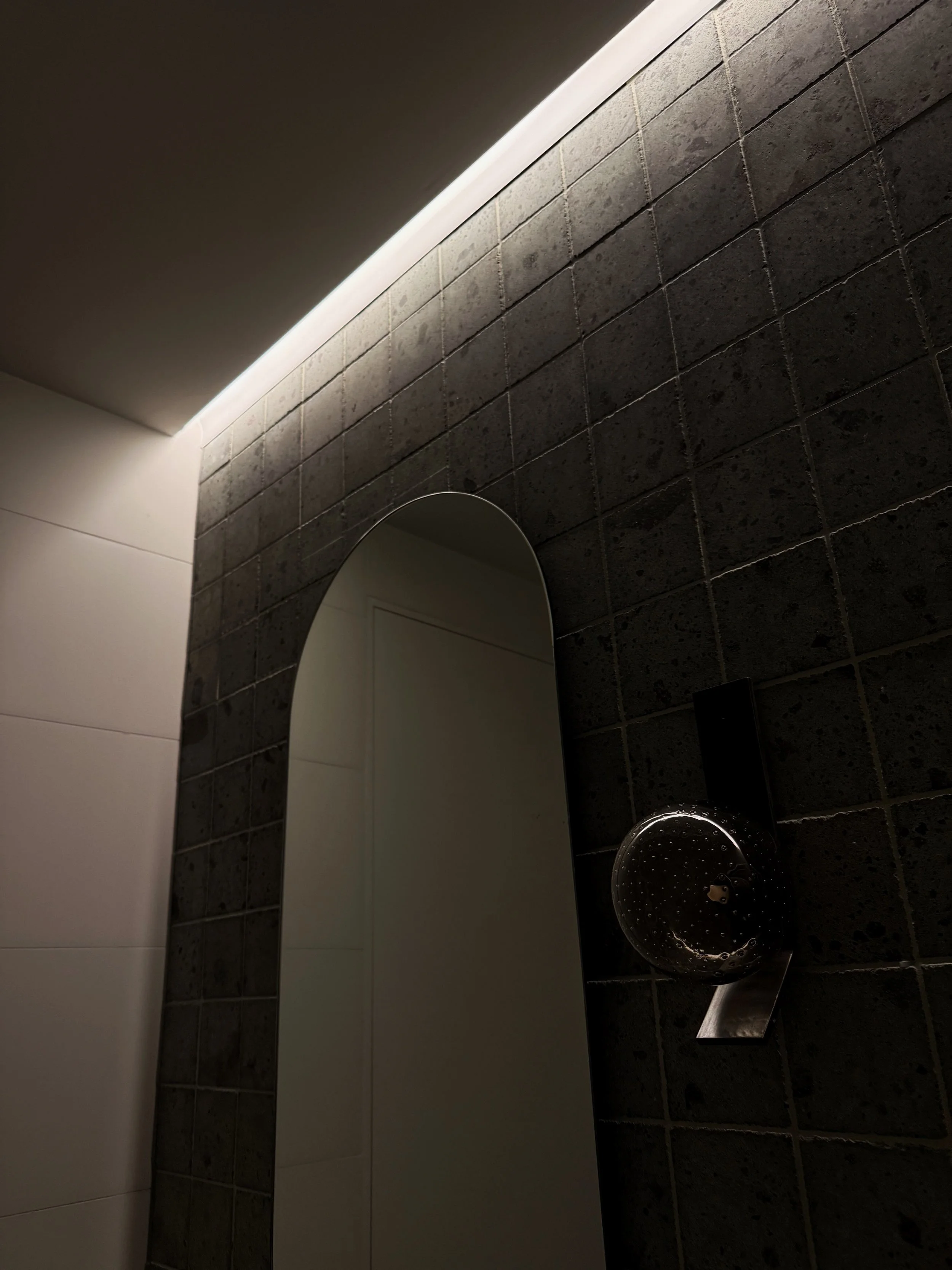 A modern bathroom with dark tiled walls, a curved mirror, and a metallic sink with a black rectangular faucet.