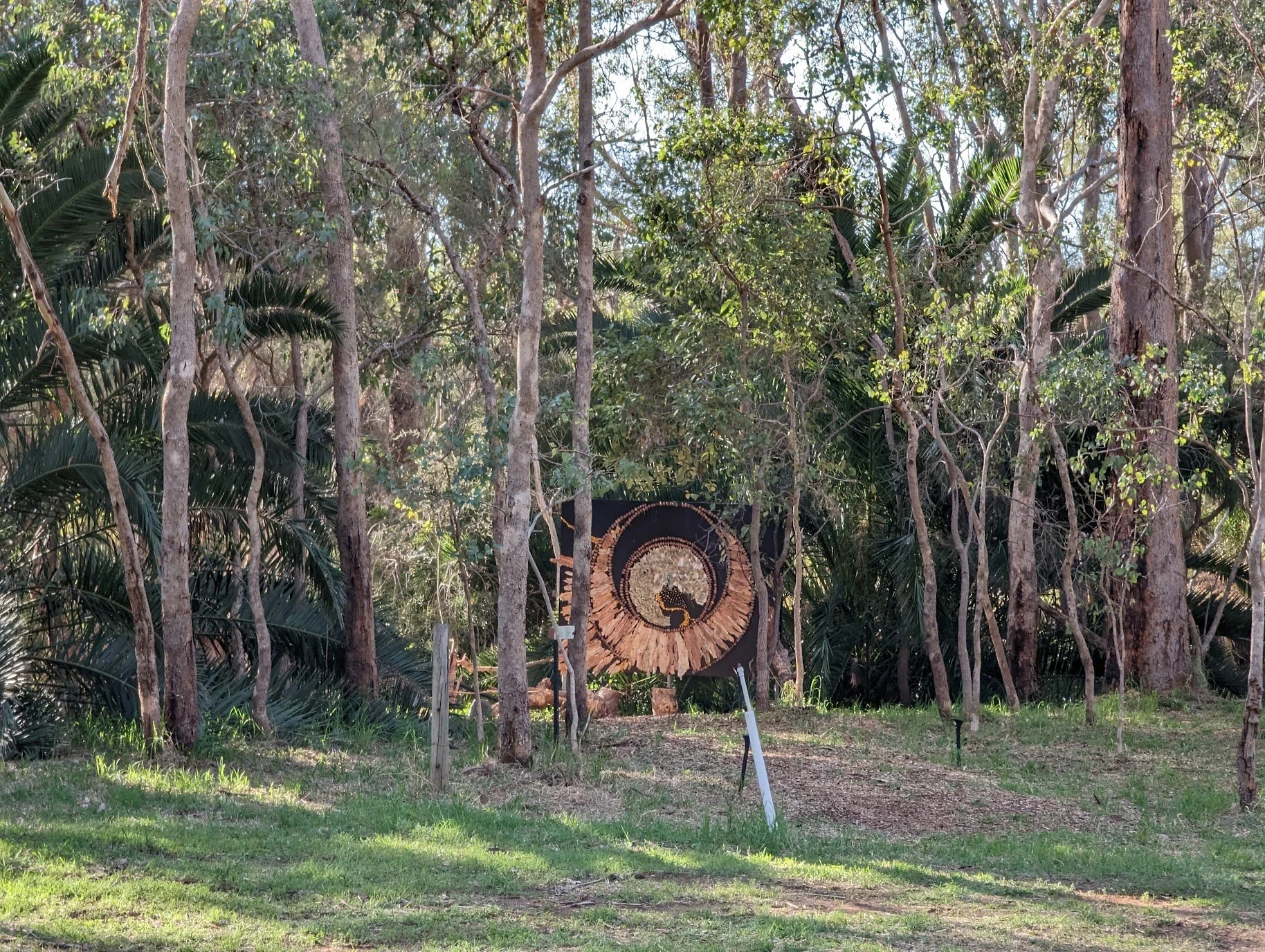 A large circular art piece made of various natural materials, including wood and stones, displayed among trees and greenery in a forested area.