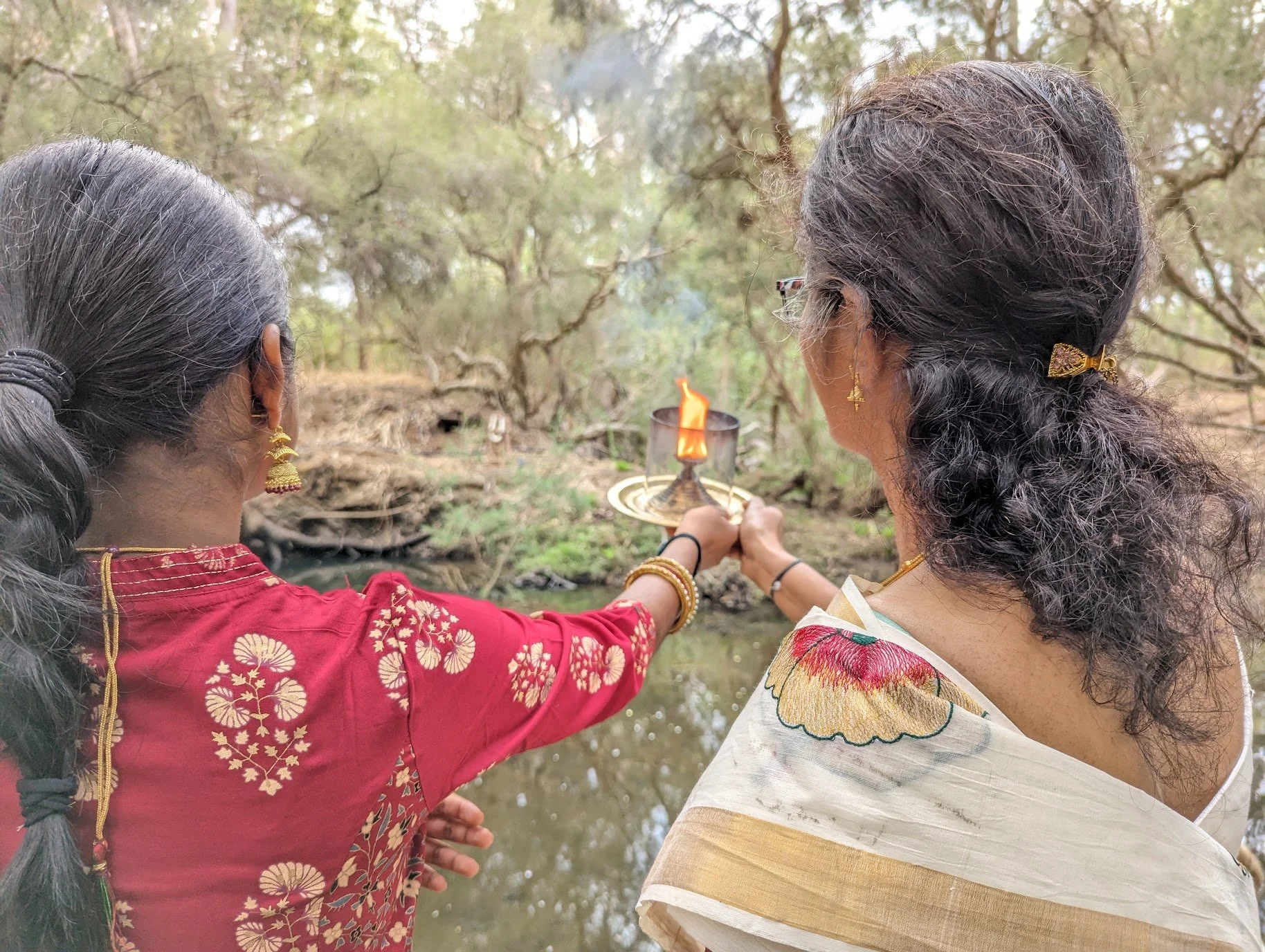 Two women standing near a river, holding a lit lamp together during a ritual or prayer, surrounded by trees.