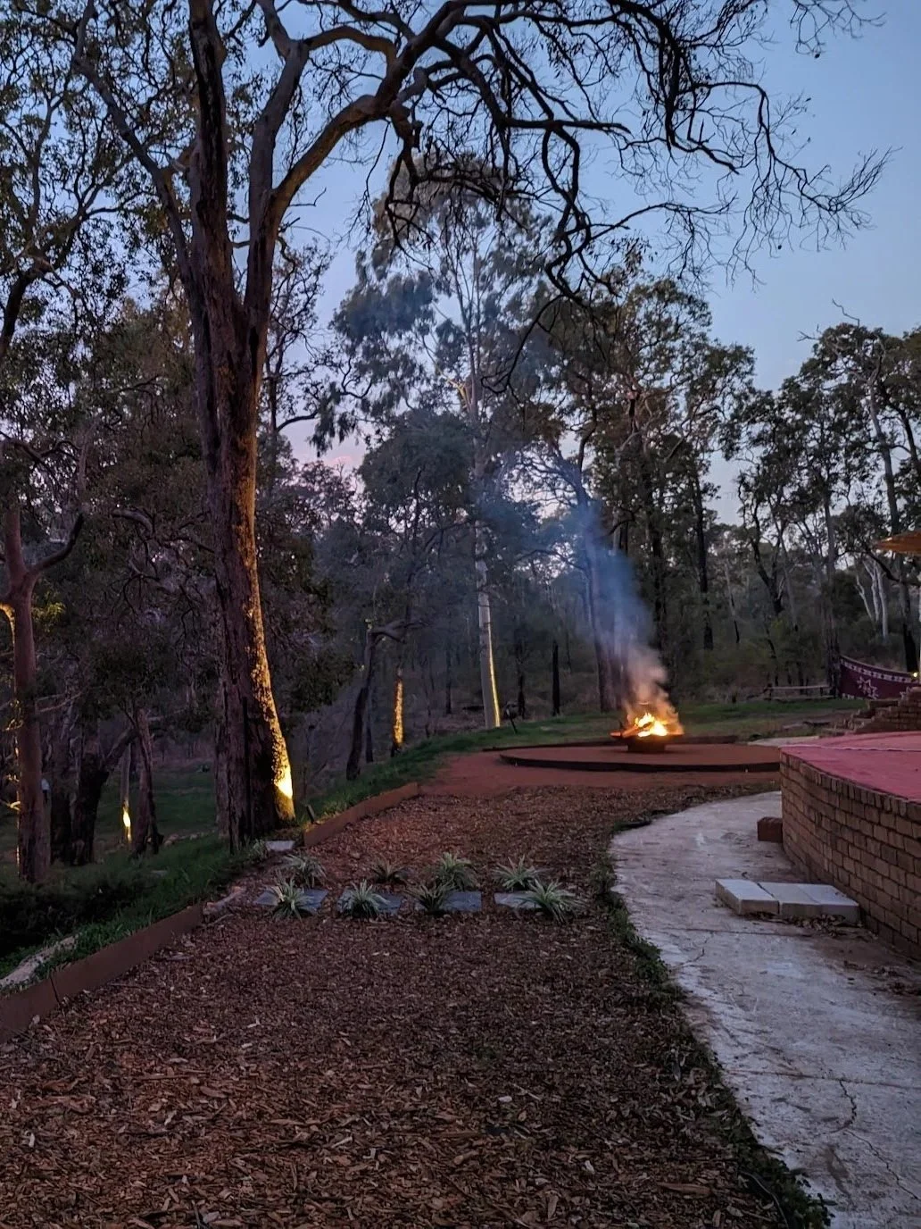A peaceful outdoor scene at dusk with trees, a fire burning in a fire pit, and a walkway curving through the landscape.