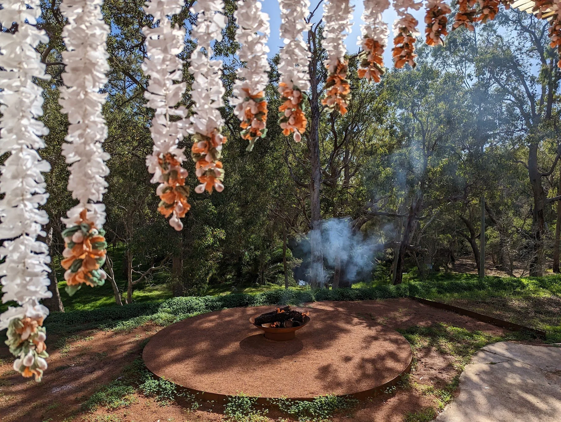 A circular fire pit on a raised platform surrounded by lush green trees and shrubs, with flowers hanging in the foreground.