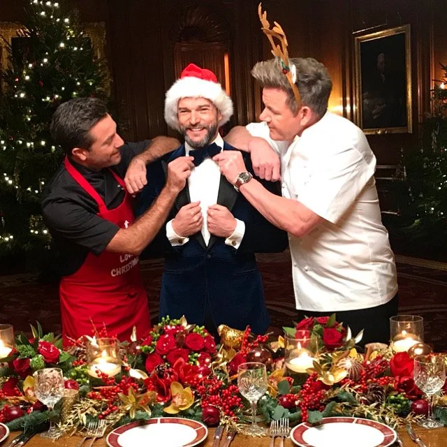 Three men celebrating Christmas at a decorated table. The man in the center is wearing a Santa hat, a tuxedo, and is being assisted by the other two men, who are adjusting his bow tie. One man is wearing a reindeer antler headband, and the other is wearing a red apron. There are Christmas-themed decorations and a Christmas tree in the background.