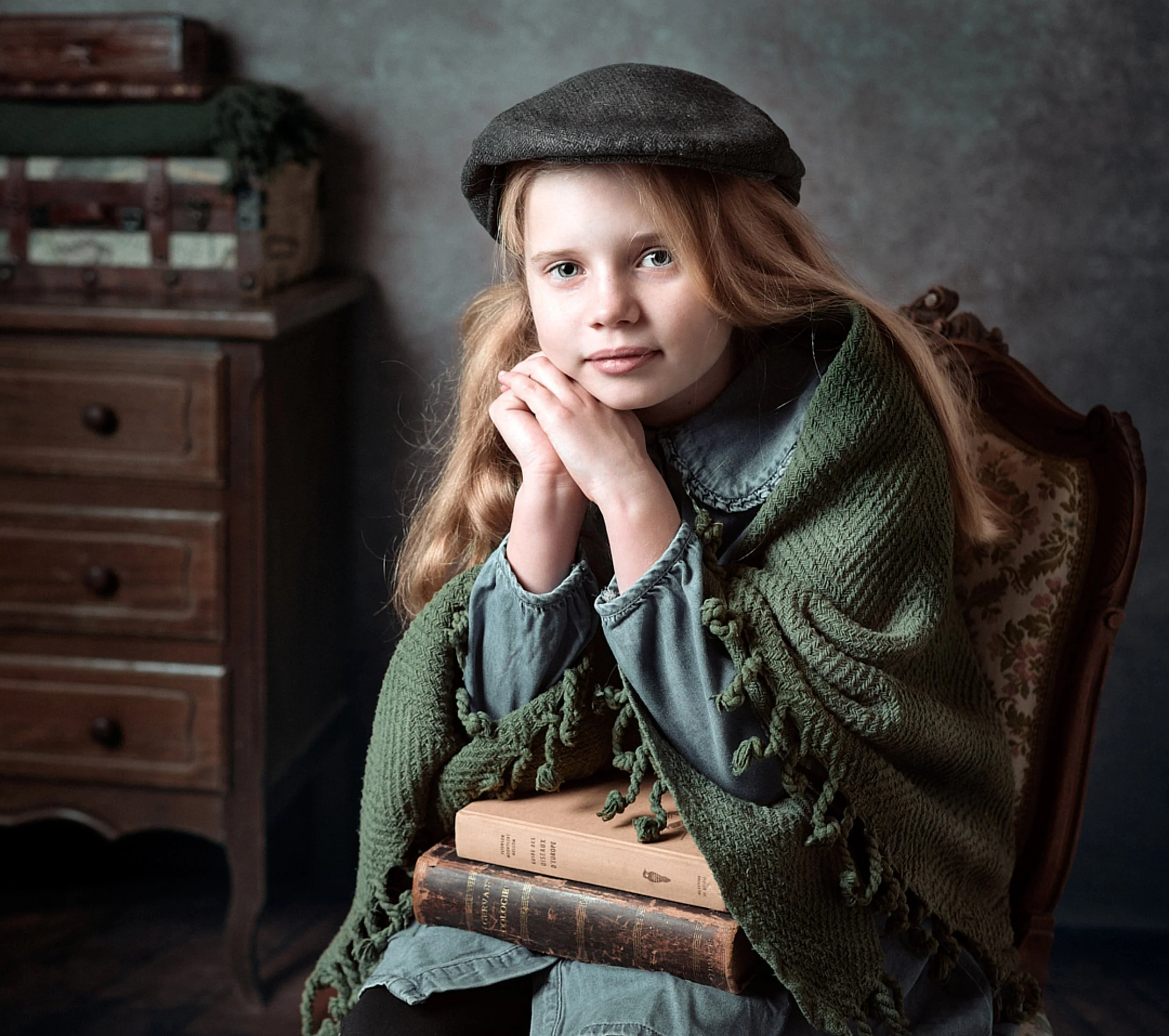 portrait fine art d'une jeune fille avec des cheveux roux, portant une casquette grise et une écharpe, assise sur une chaise en bois recouverte d'une couverture verte, tenant deux livres, avec un fond sombre.