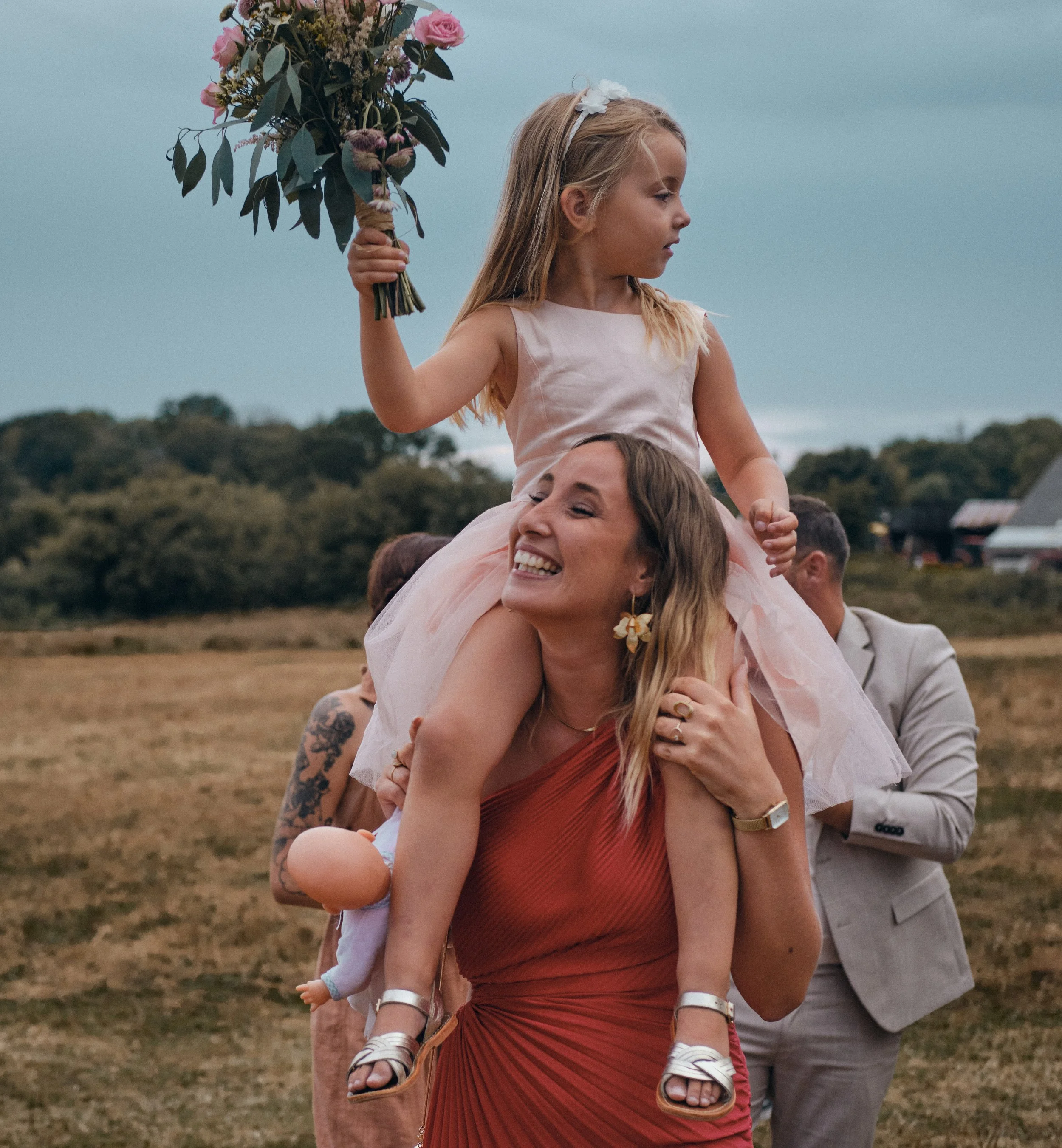 Lors d'un mariage dans le Jura près de Champagnole Une jeune fille assise sur les épaules d'une femme, tenant un bouquet de fleurs, lors d'une célébration extérieure avec un groupe de personnes dont un homme portant un costume à tortues, un paysage a