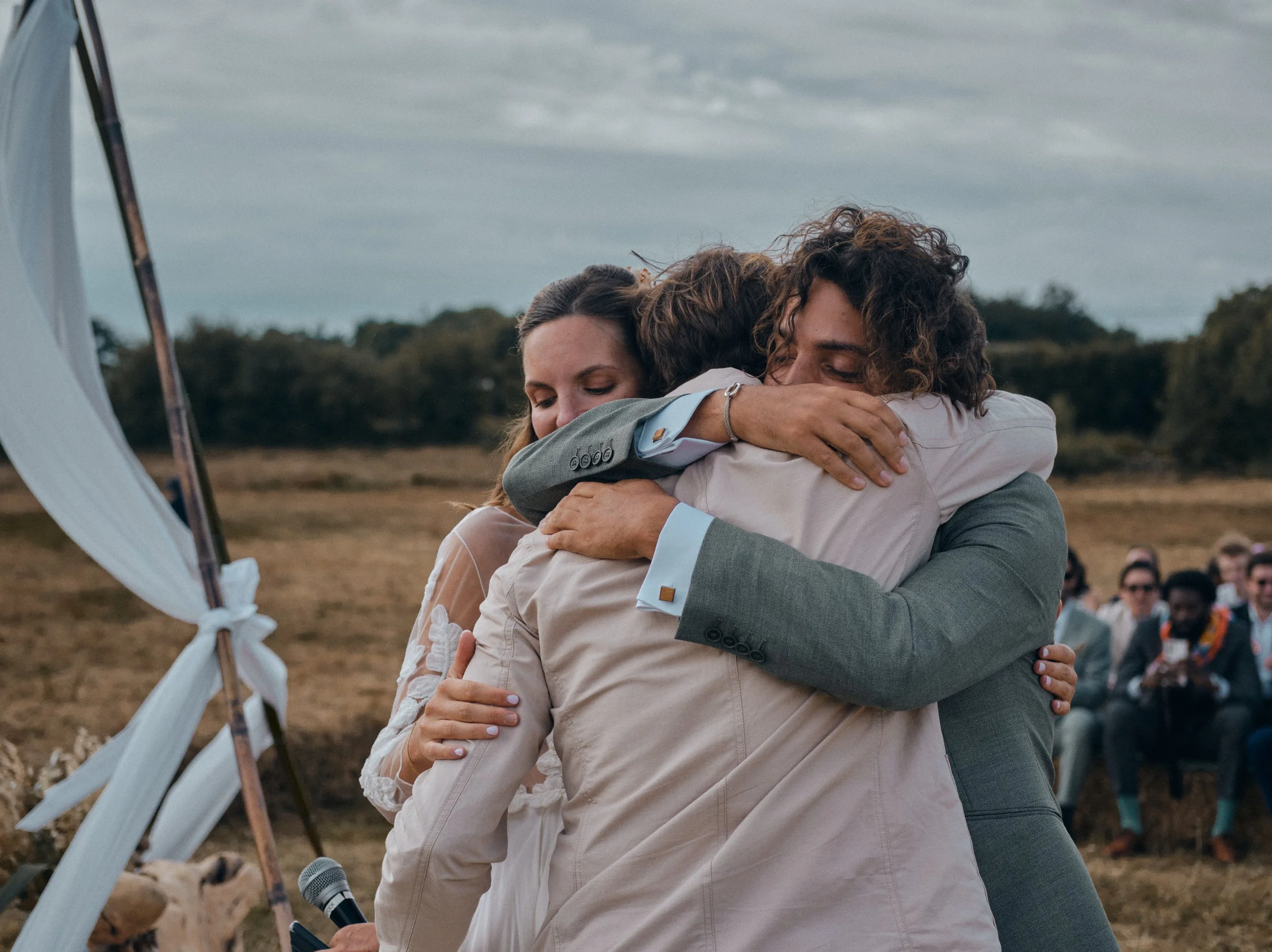 Deux femmes en costume élégant s'embrassent lors d'une cérémonie en plein air avec un groupe de spectateurs en arrière-plan.