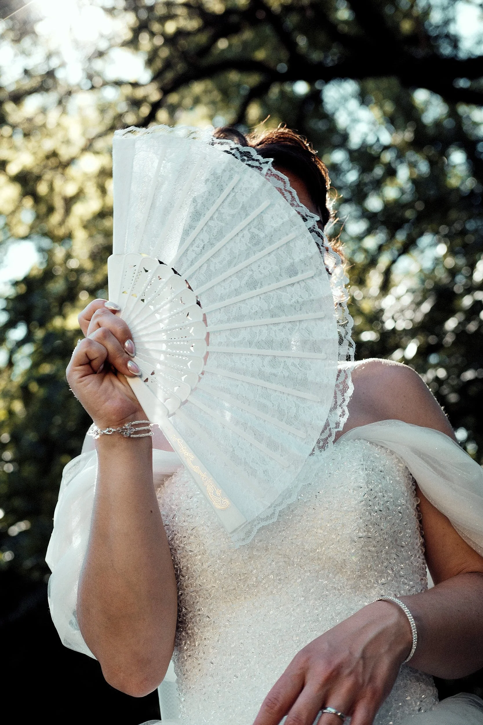 Personne en robe de mariée tenant un éventail blanc en dentelle devant le visage, avec un arrière-plan d'arbres et de lumière naturelle, lors d'un mariage près de Champagnole dans le Jura.