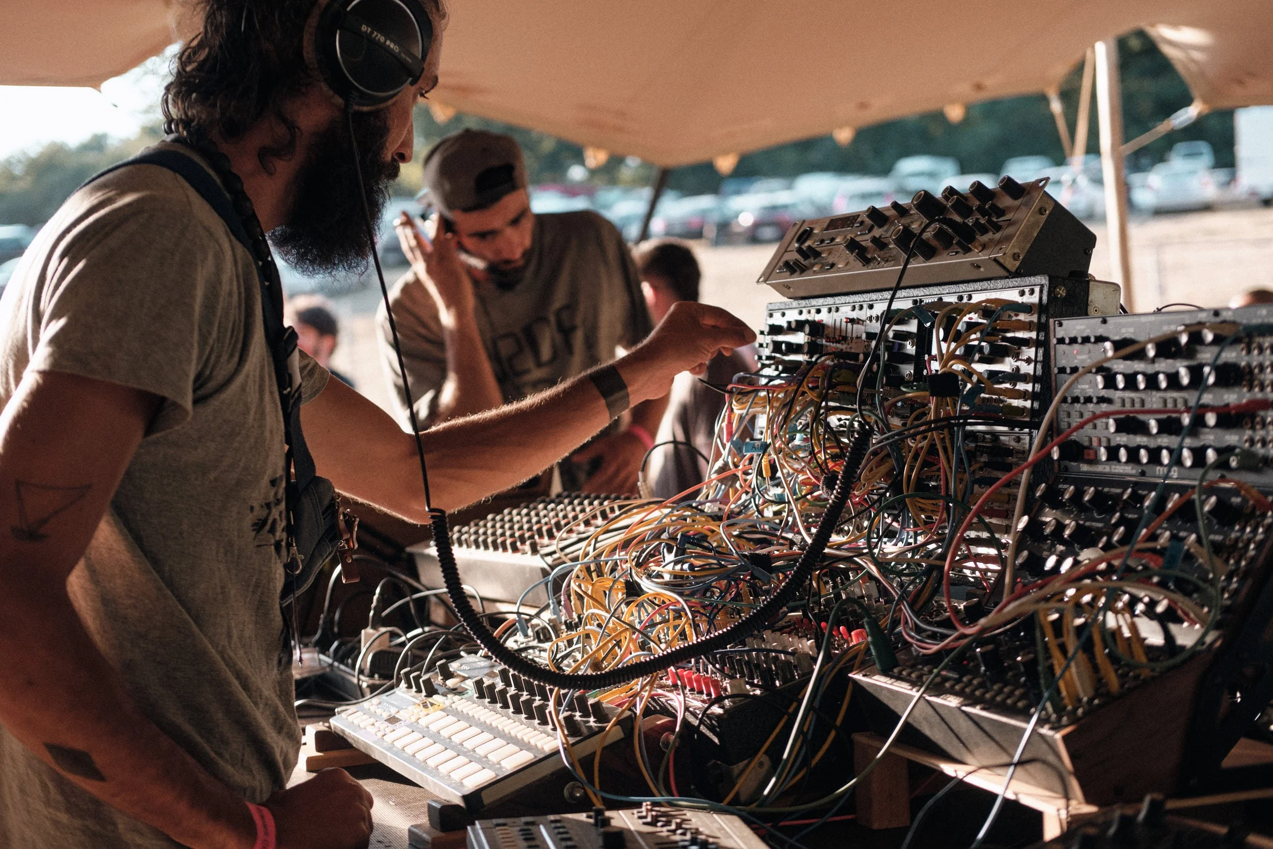 Deux musiciens jouent avec un synthétiseur et un séquenceur dans un espace en plein air sous une tente, avec des câbles et du matériel électronique dispersés.