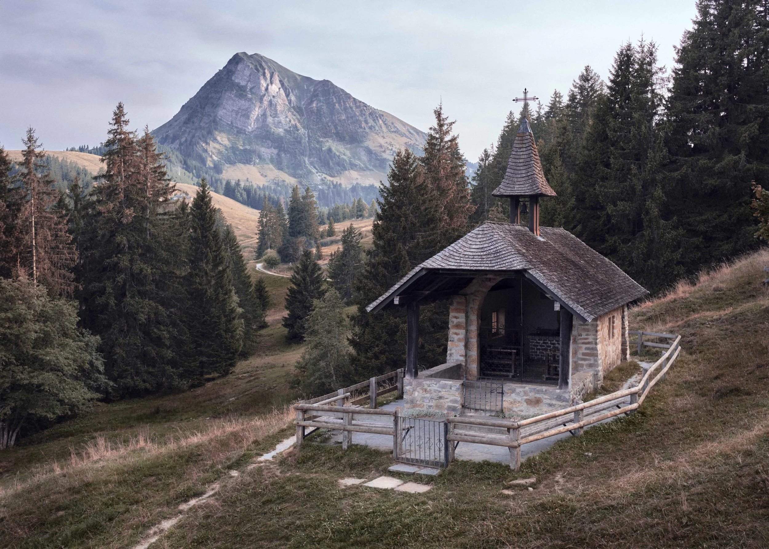 Petite chapelle en pierre avec un toit en tuiles en bois, entourée d'une clôture en bois, située dans une région montagneuse avec des arbres et une montagne en arrière-plan.