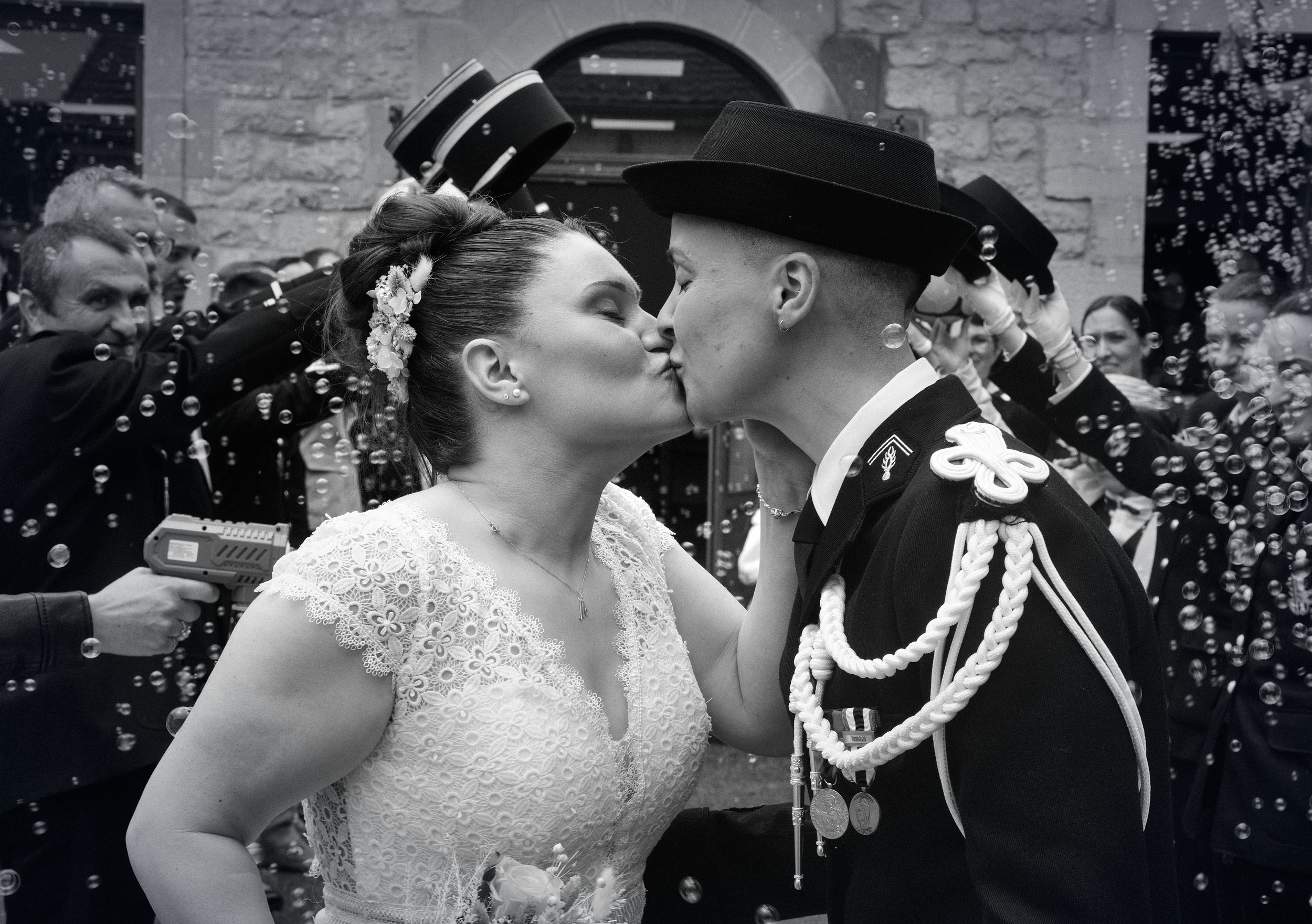 Un couple habillé pour un mariage, la femme en robe blanche tenant un bouquet de fleurs roses et le homme en uniforme militaire, se tenant côte à côte et souriant.