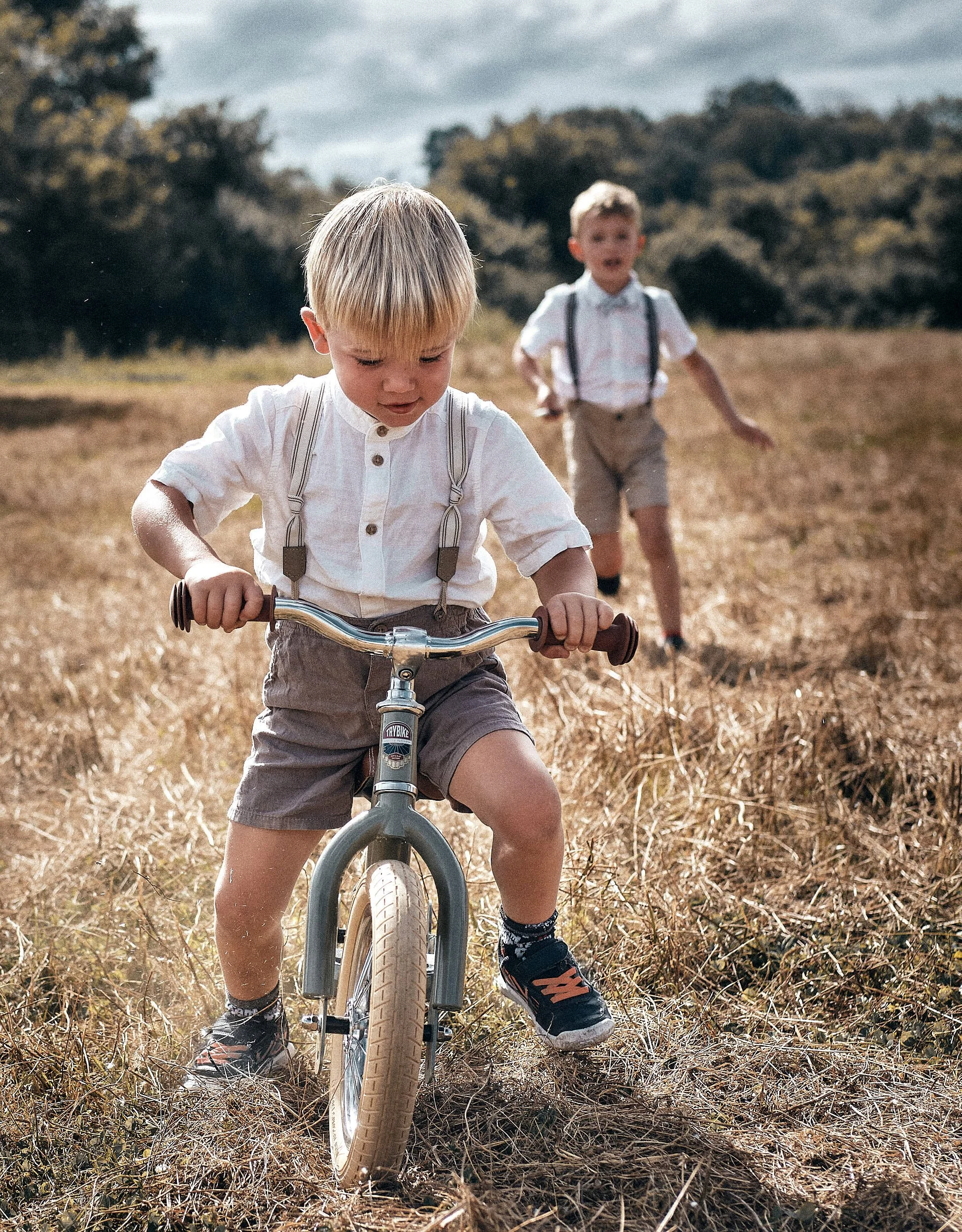 Lors d'un mariage deux jeunes garçons jouent dans un champ sec dans le Jura près de Champagnole, l'un sur un vélo, l'autre court derrière, avec des arbres en arrière-plan et un ciel nuageux.