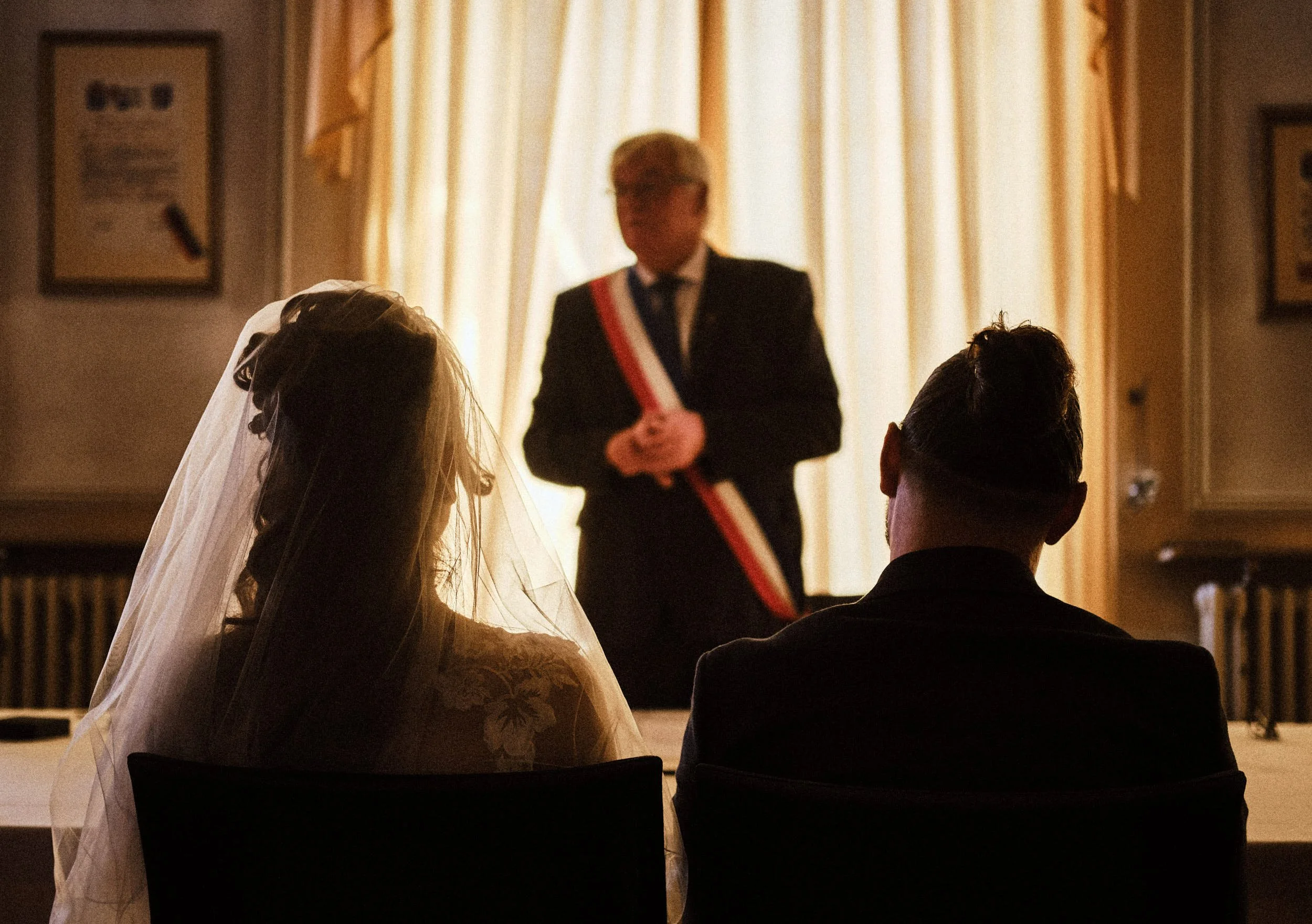 Photographie d'une cérémonie à la mairie de Champagnole dans le Jura avec une femme en robe de mariée et plusieurs personnes assises. Le maire en uniforme avec une écharpe tricolore se tient debout en arrière-plan pour célébrer leur union