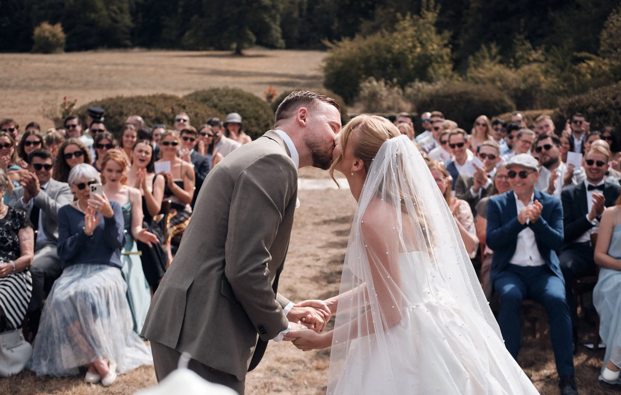 Photographie d'un couple de mariés échangeant un baiser lors d'une cérémonie de mariage en plein air au Château de la Muyre dans le jura, entourés d'invités assis et applaudissant dans un champ avec des arbres en arrière-plan.