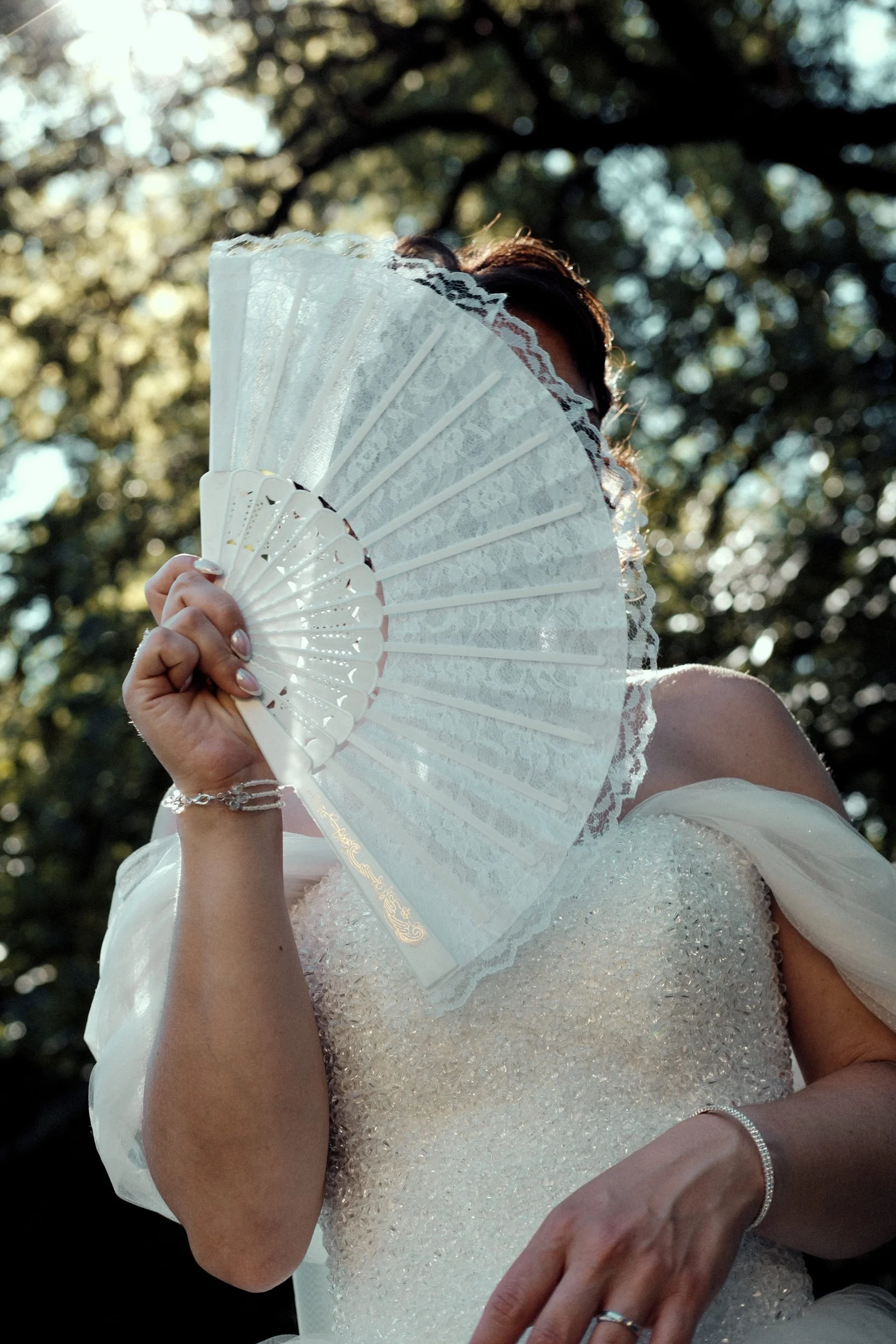 Personne en robe de mariée tenant un éventail blanc en dentelle devant le visage, avec un arrière-plan d'arbres et de lumière naturelle, lors d'un mariage près de Champagnole dans le Jura.