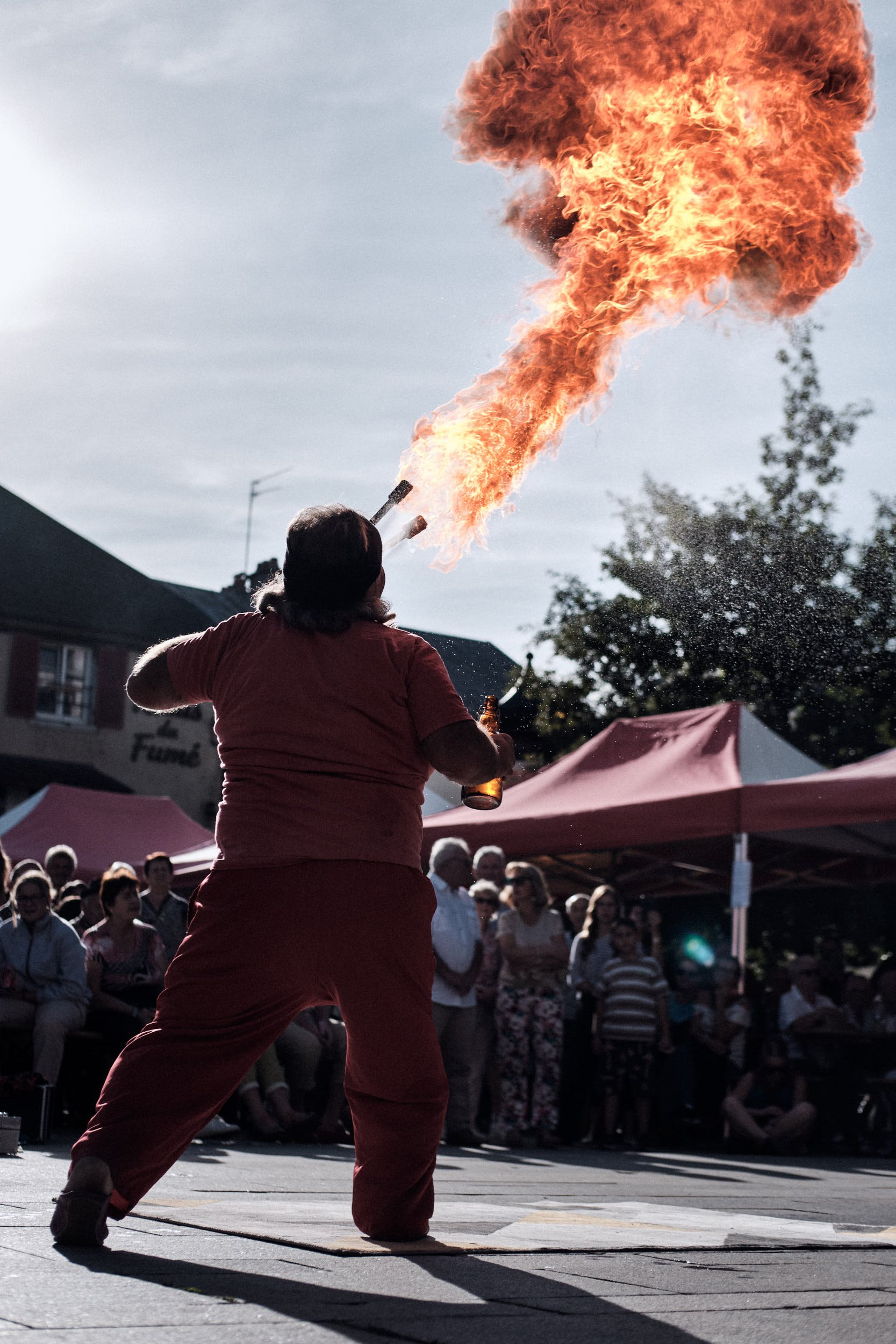 Une femme en rouge lance du feu lors d'une performance publique devant une foule assise et debout, sous un ciel nuageux, avec des tentes autour.