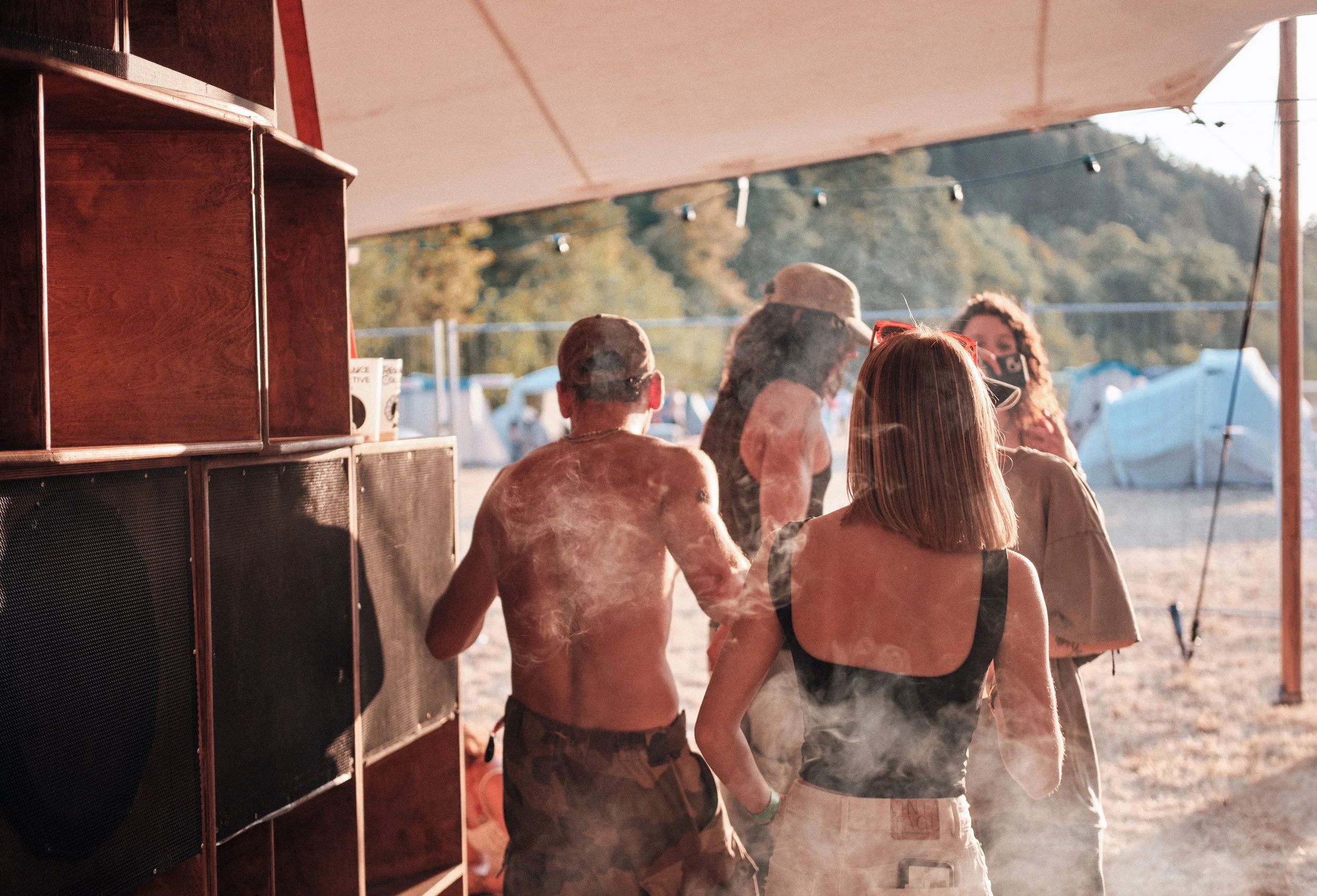 Groupe de personnes à un festival en plein air, vue depuis l'intérieur d'une tente ou d'une cabine, avec des arbres en arrière-plan.