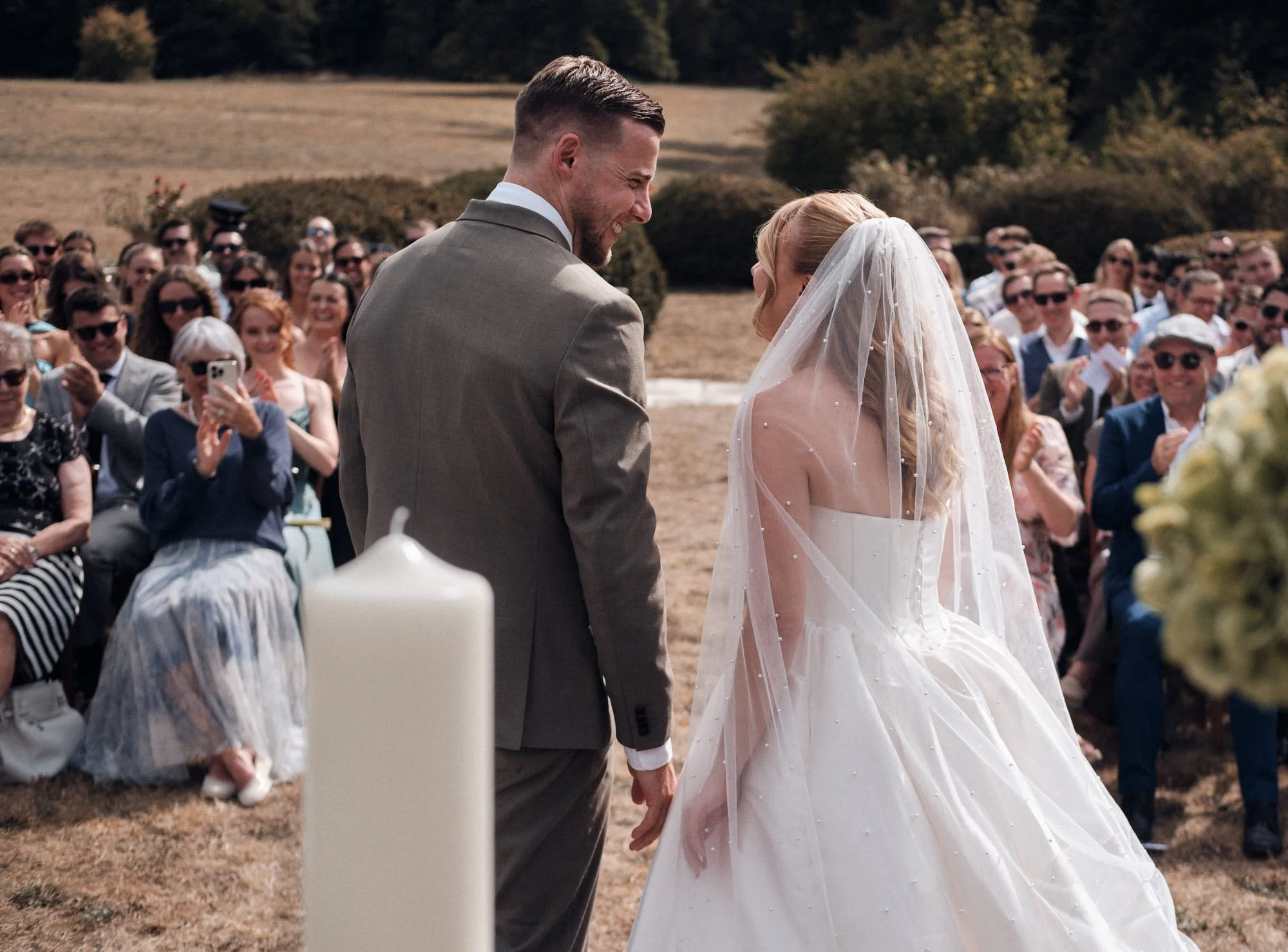 Un couple de mariés échangeant un baiser lors d'une cérémonie en plein air, entourés d'invités qui prennent des photos, avec un fond de campagne ensoleillée.