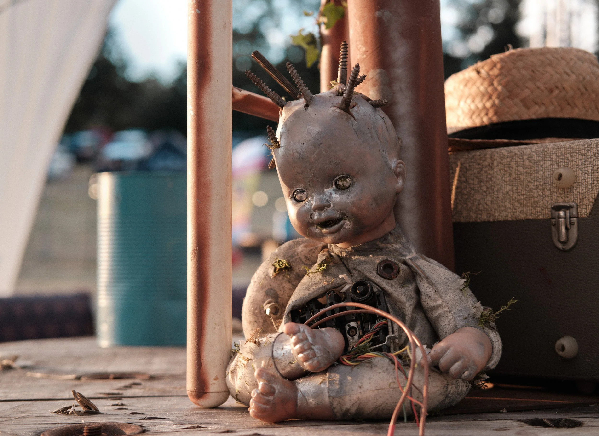 Une poupée en plastique dégradée avec des vis enfoncées dans la tête, assemblage d'objets de récupération avec des fils électriques visibles, posée sur un sol en bois.