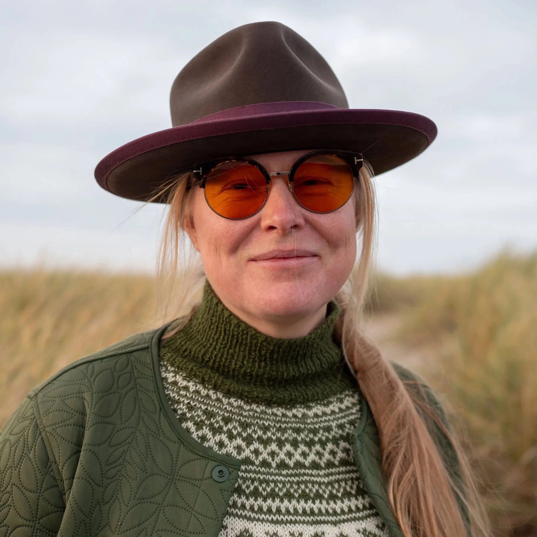 Kvinde med bredbrimmet hat, runde briller med orange glas, grøn sweater og grøn jakke, står udendørs med græs i baggrunden.