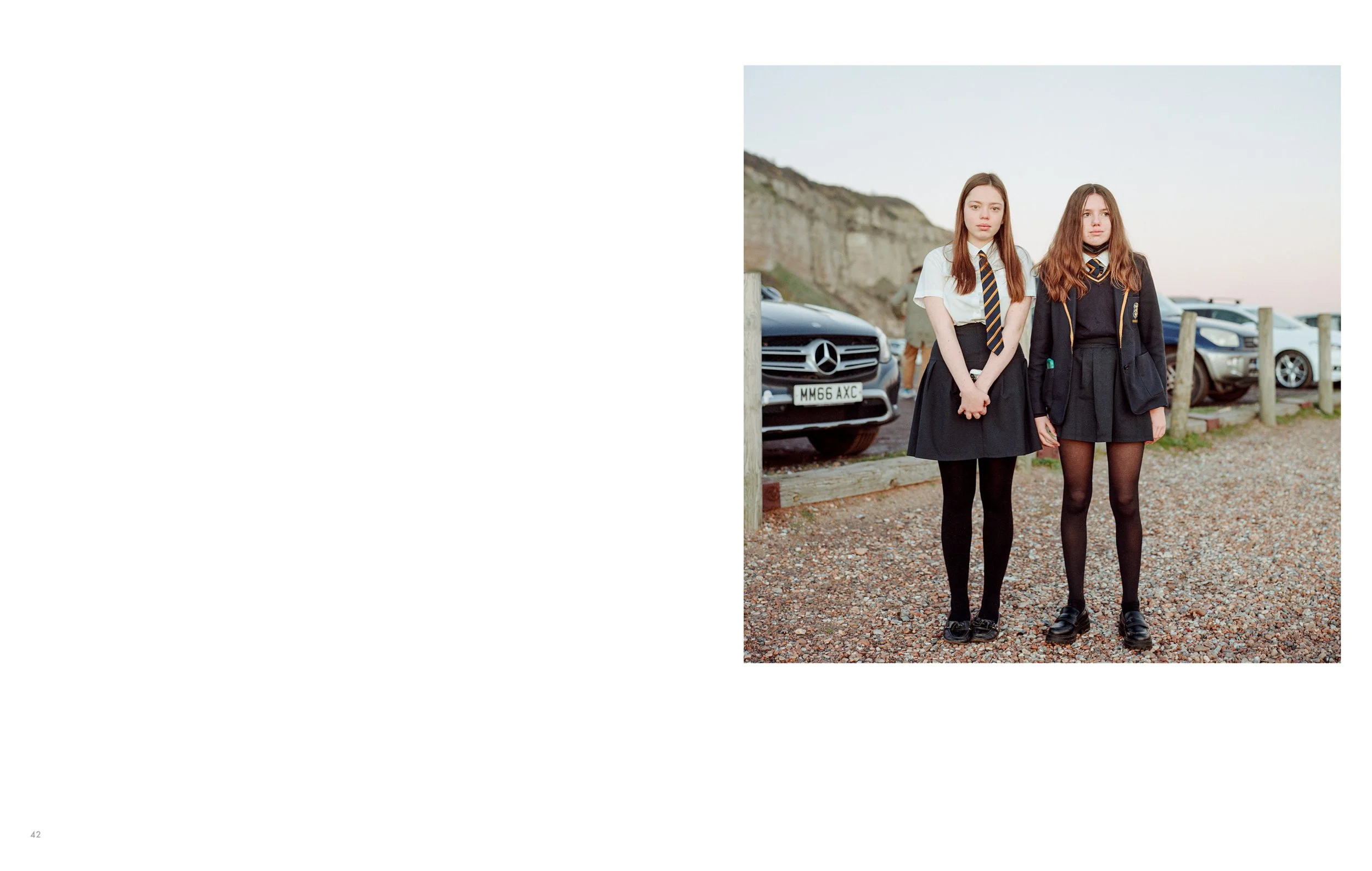 Two teenage girls dressed in school uniforms standing outdoors near parked cars, with a rocky hillside in the background during the late afternoon or early evening.
