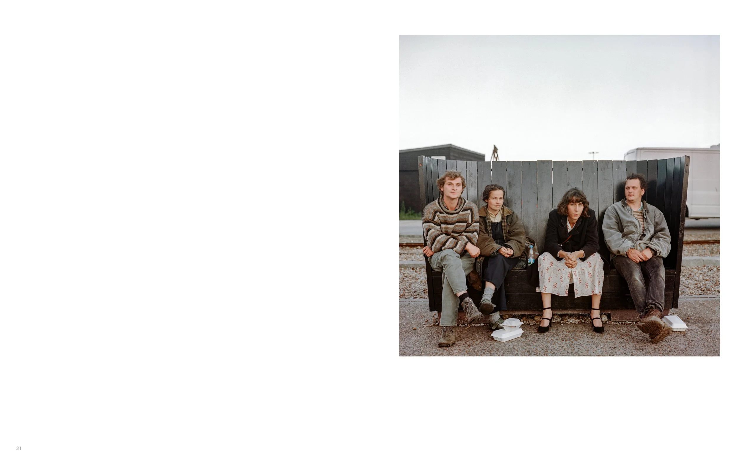 Four young adults sitting on a bench in front of a black wooden fence outdoors, with overcast sky, casual clothing, and some items at their feet.
