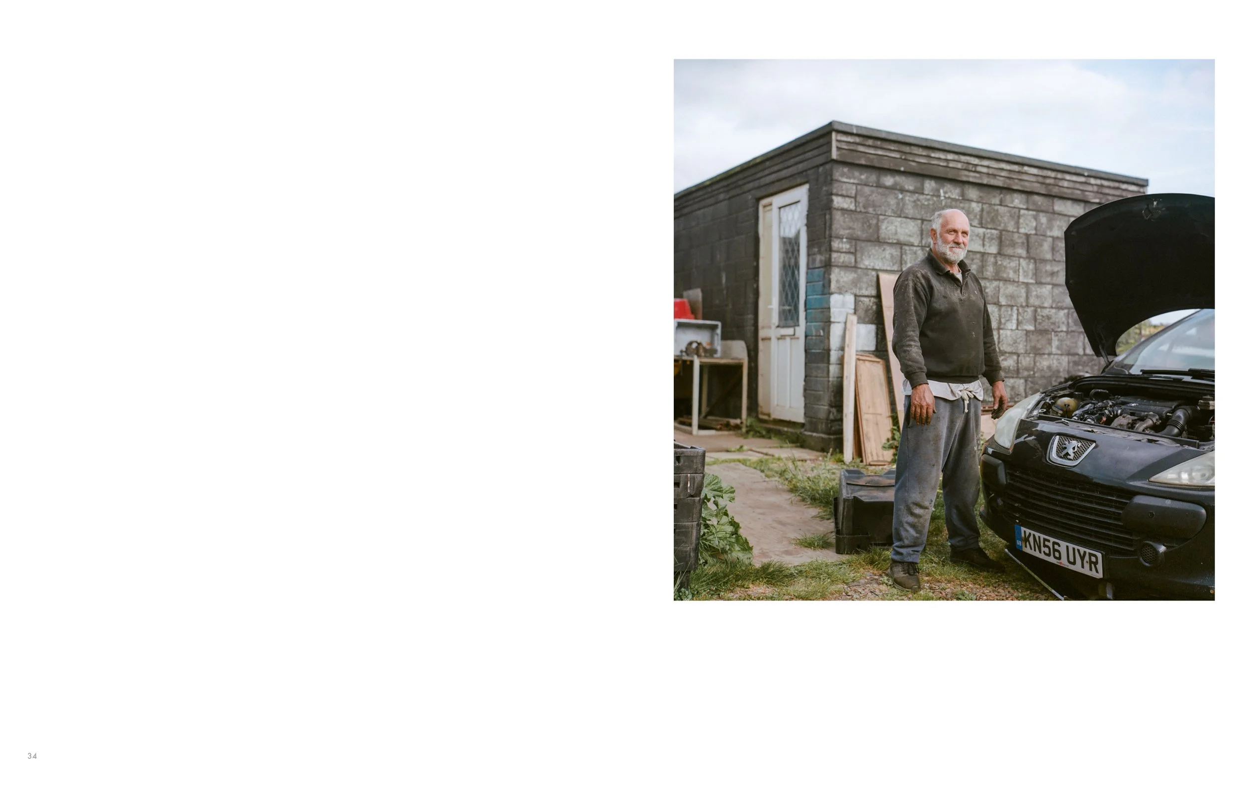 An elderly man with a beard standing next to a black car with its hood open, in front of a gray brick building. The man appears to be working on the car.