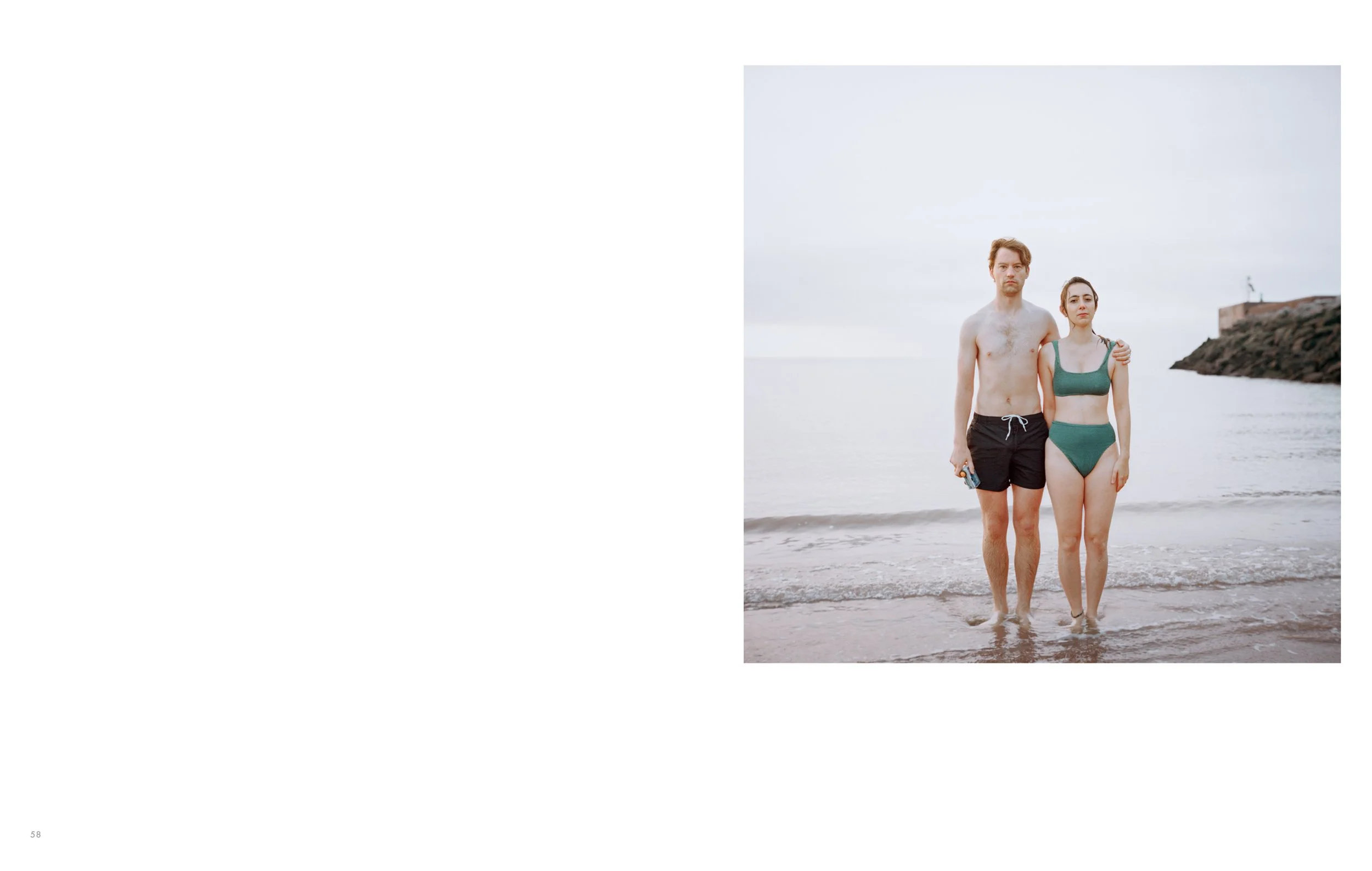 A young man and woman standing close together on a beach with their feet in the water, facing the camera. The man is shirtless, wearing black shorts, and the woman is in a teal swimsuit. The sky is cloudy, and a rocky shoreline with a building and a 