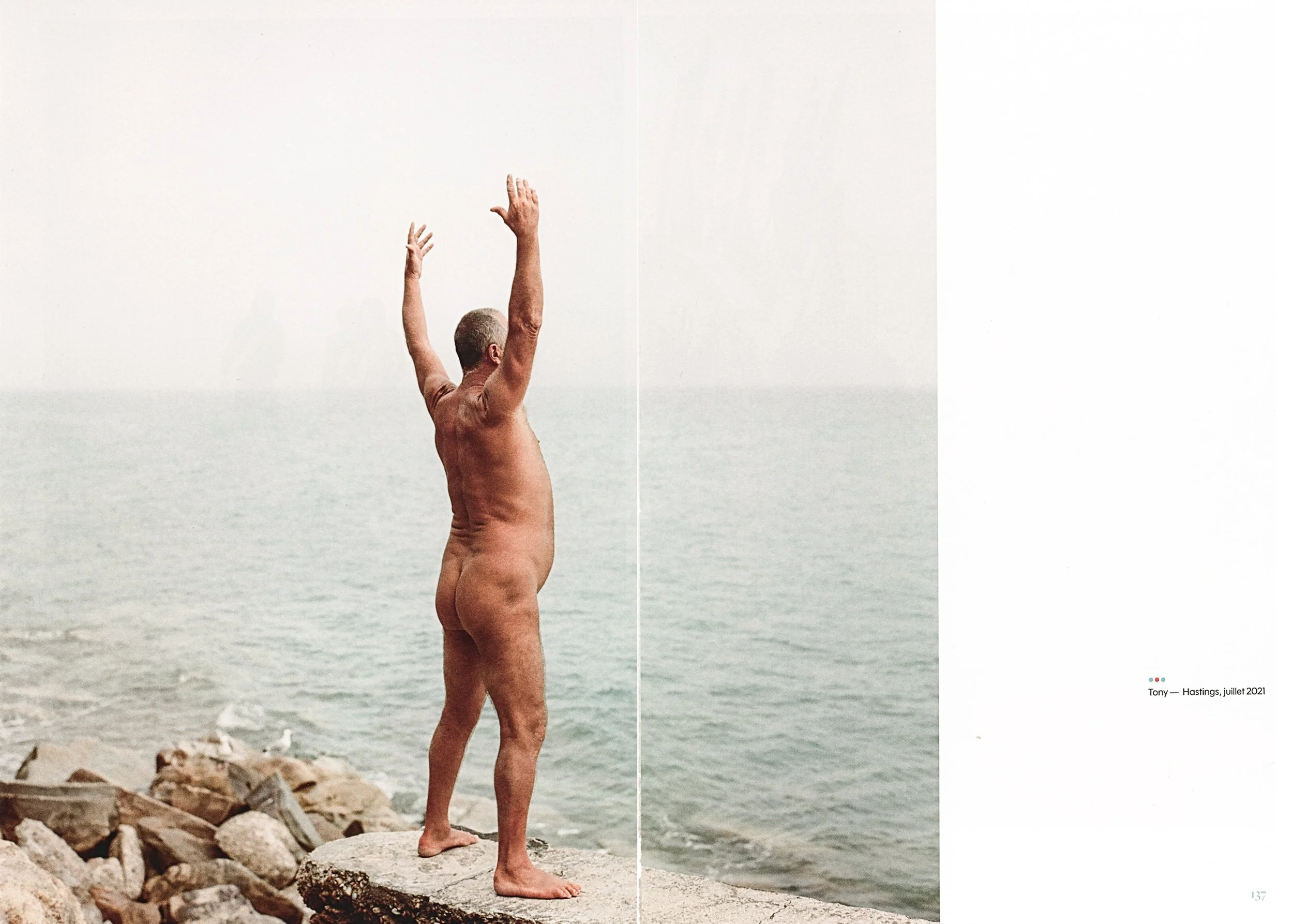 A person standing nude on a rocky seashore with arms raised, facing the ocean.