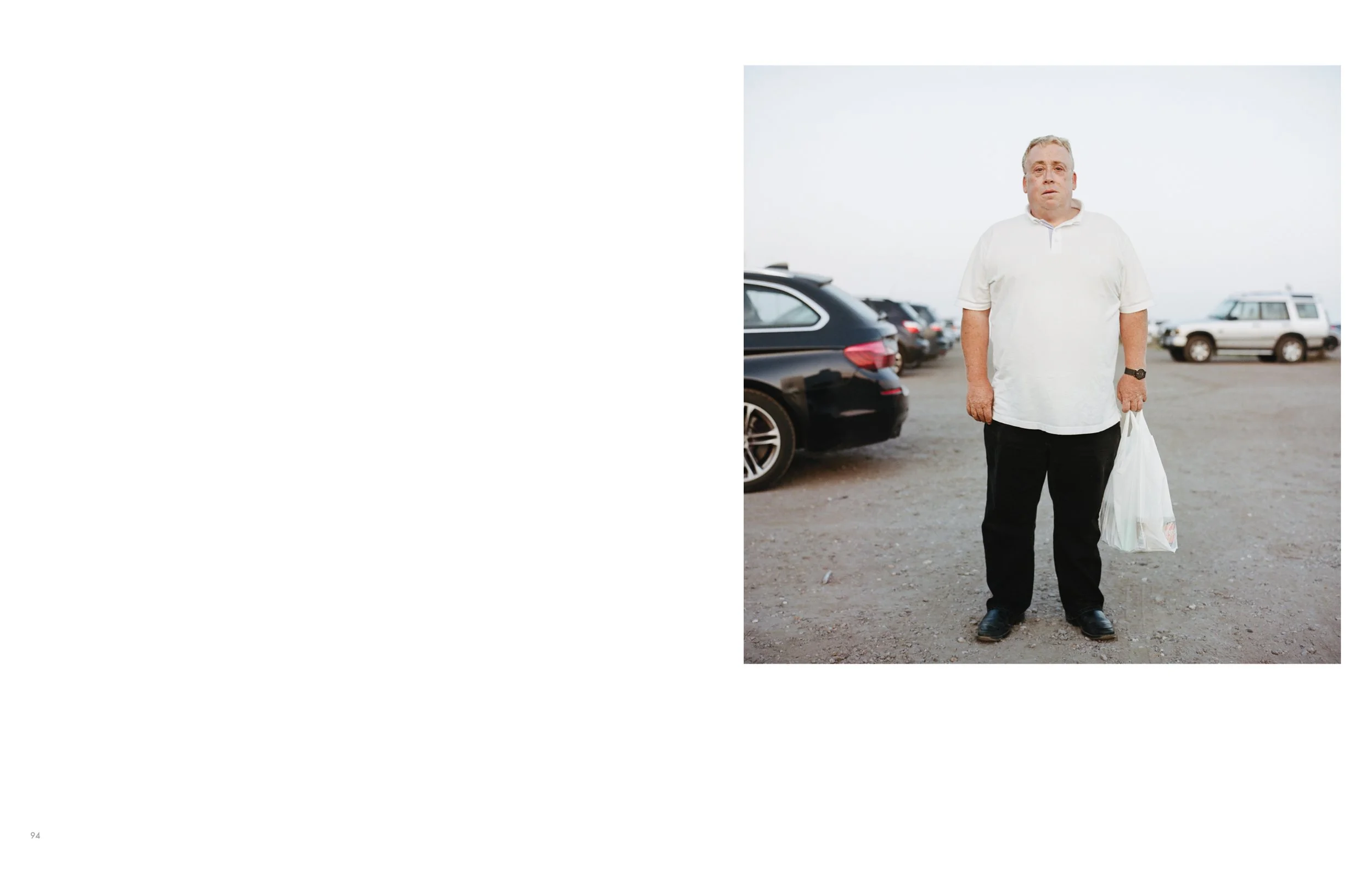 An older man with gray hair and glasses stands outdoors on a parking lot, holding a plastic grocery bag in his left hand, wearing a white polo shirt, black pants, and a wristwatch, with cars parked in the background under an overcast sky.