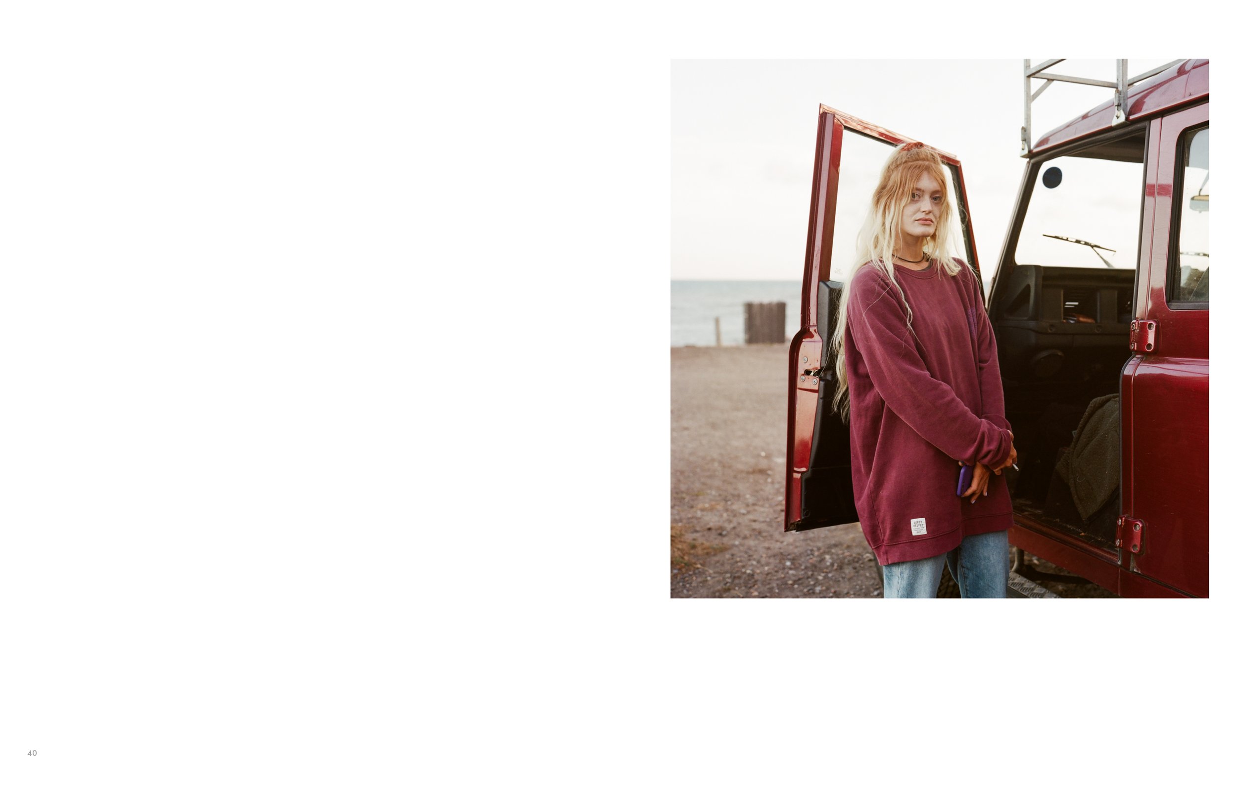 A young woman with long blonde hair standing next to a red vehicle at the beach, wearing a maroon oversized sweatshirt and holding a phone.